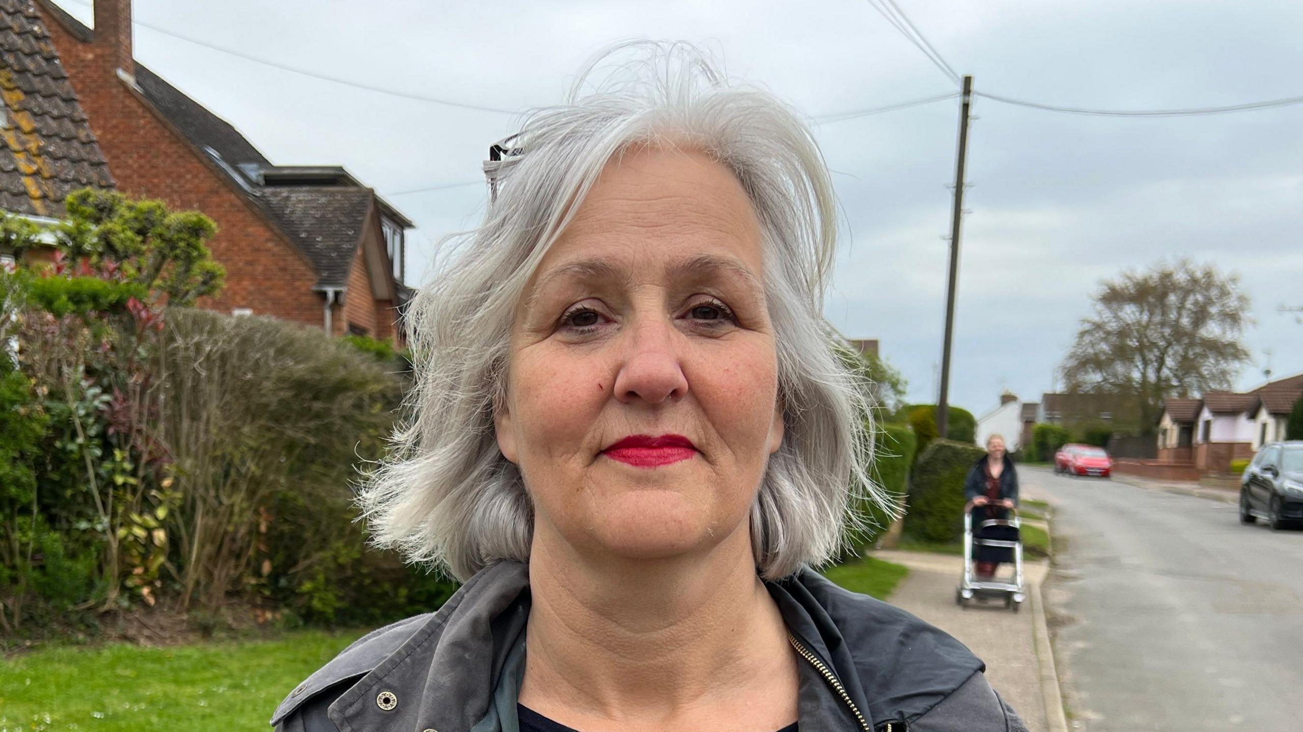 A smiling woman looks straight at the camera while standing on a pavement. She is wearing a grey coat.