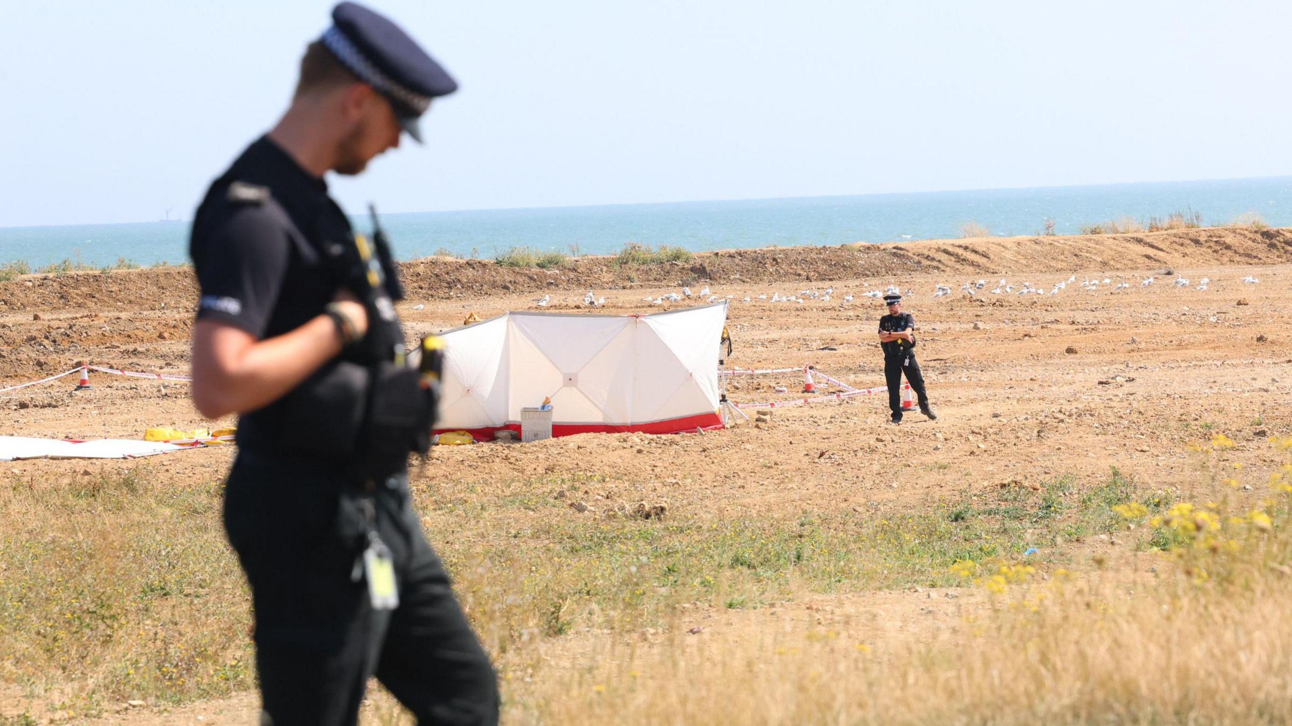 A police officer stands in the foreground. In the background is a white police tent just in front of the beach. A police officer stands next to it.