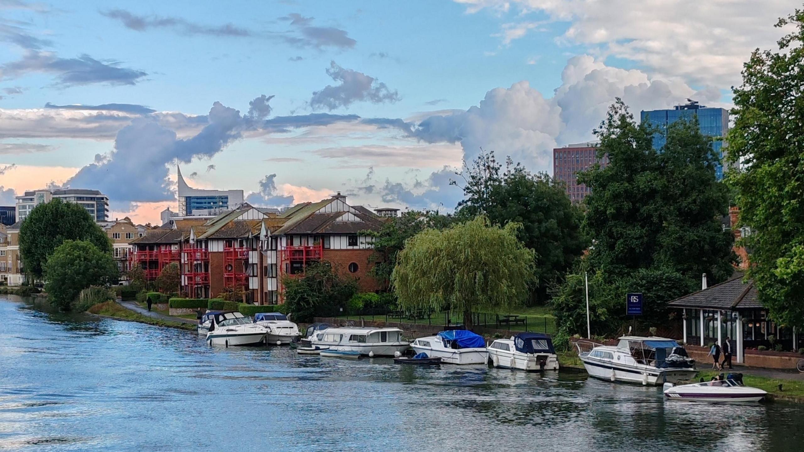 Five small white boats are moored in the water next to a path, with several tall buildings making up the skyline under a mostly blue sky with several white clouds. 