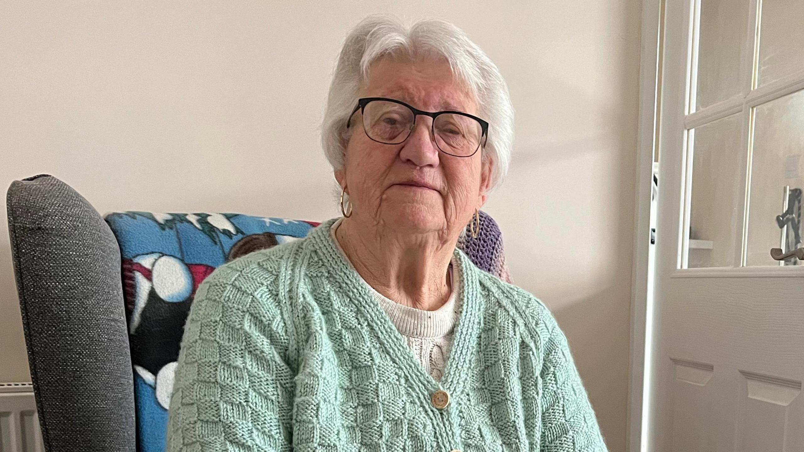 An elderly woman with white hair wearing glasses and looking at the camera. She is wearing a mint green cardigan and is sitting on a grey chair