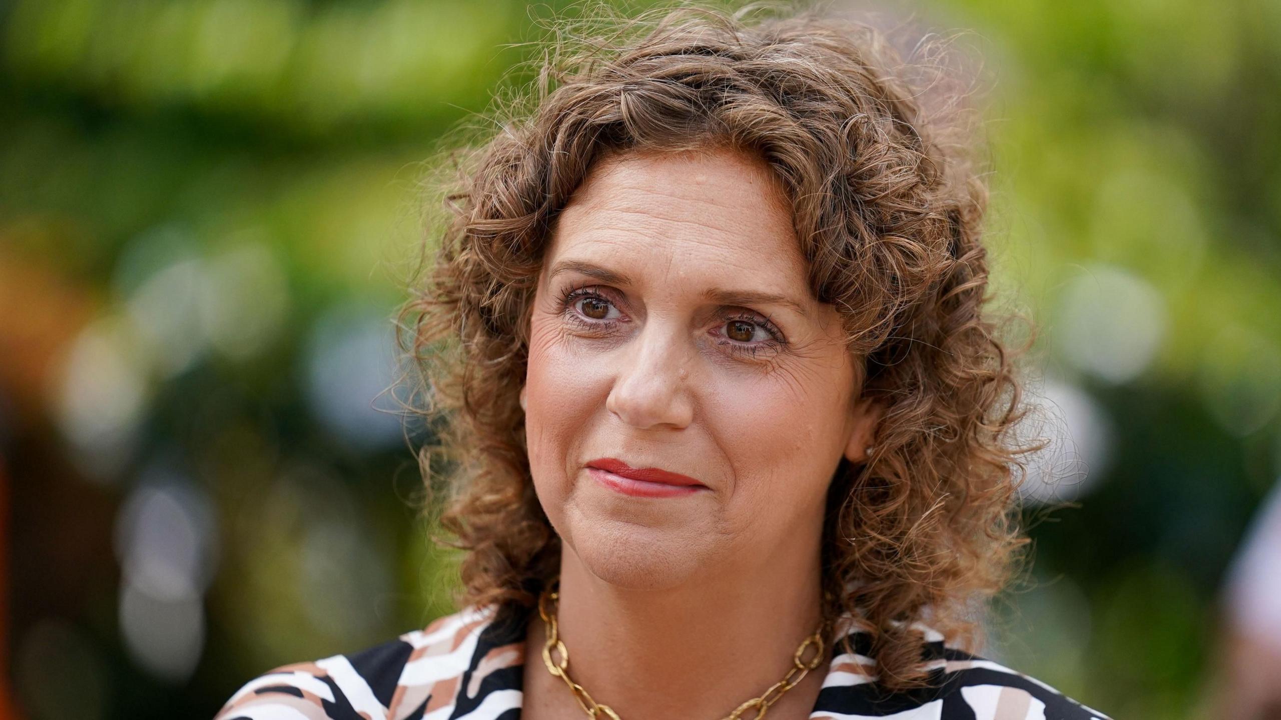 Hannah Ingram-Moore, Captain Tom's daughter, is photographed in Captain Tom's Garden. She is wearing a gold necklace and a patterned blouse which is black, white and orange.