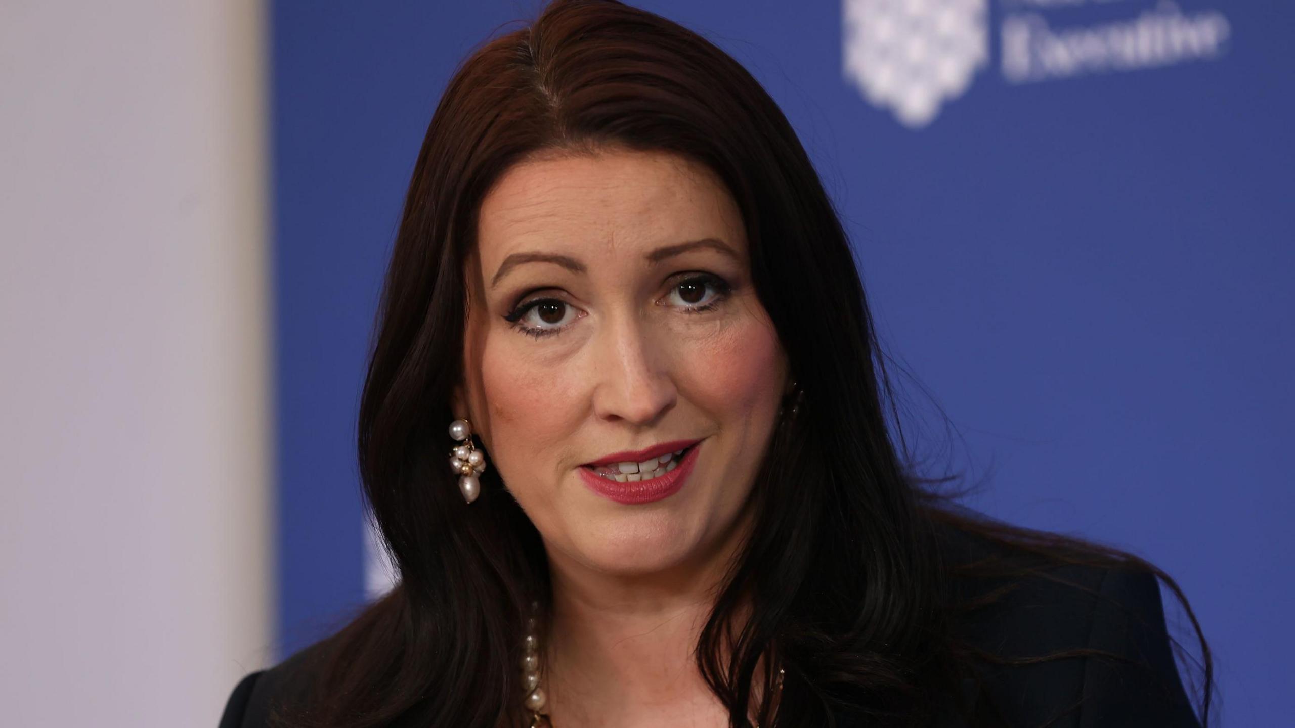 Northern Ireland Deputy First Minister Emma-Little Pengelly is speaking during a press conference. We can see the tops of her shoulders and head. She has shoulder-length dark hair and is wearing a pearl necklace and pearl earrings. She is wearing a dark jacket. In the background is a blue board with the logo of the Northern Ireland Executive