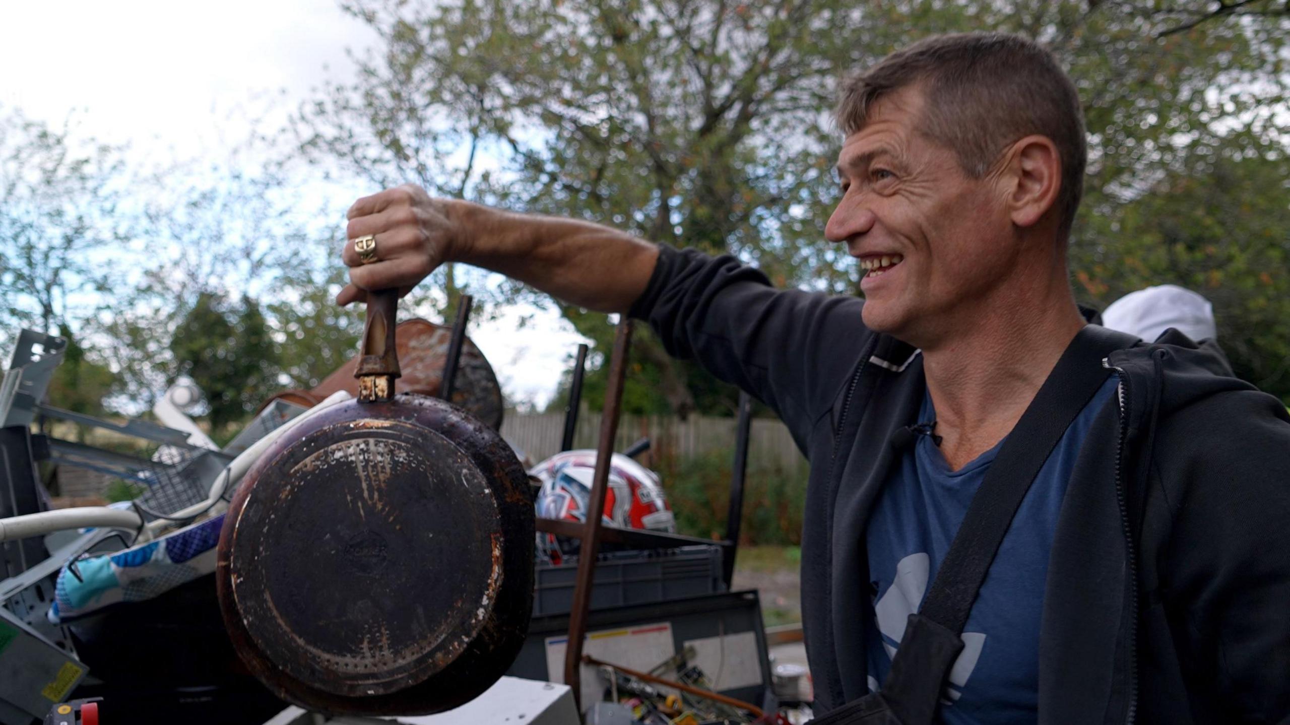 A smiling man holding up a frying pan that has been left out as rubbish. He has short, brown hair and is wearing a blue jacket and a blue t-shirt. 