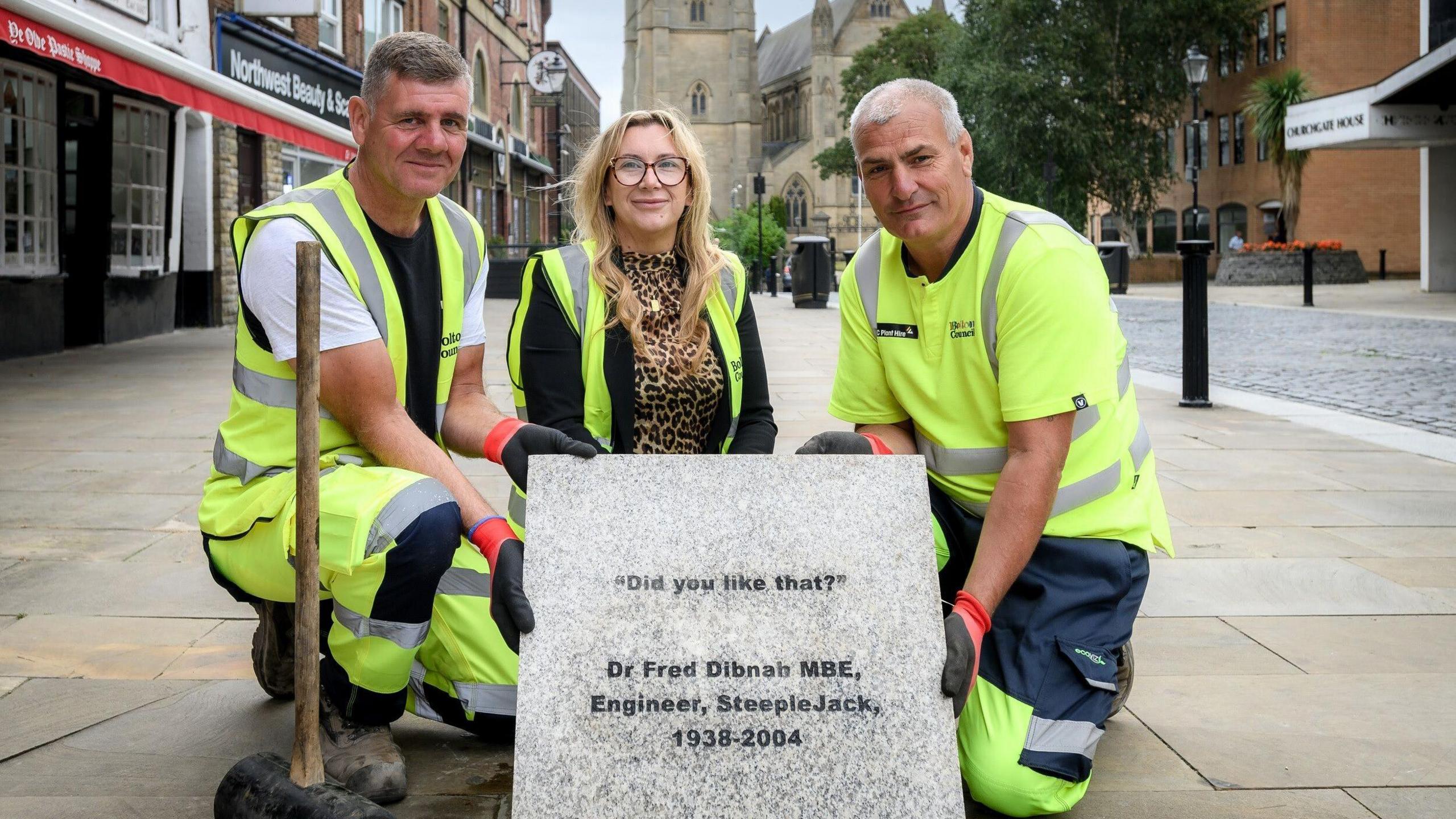 Two workmen and a female councillor all hold up the new commemorative stone to Fred Dibnah. It reads 'Did you like that?'. They are crouched down alongside the stone in Bolton town centre.