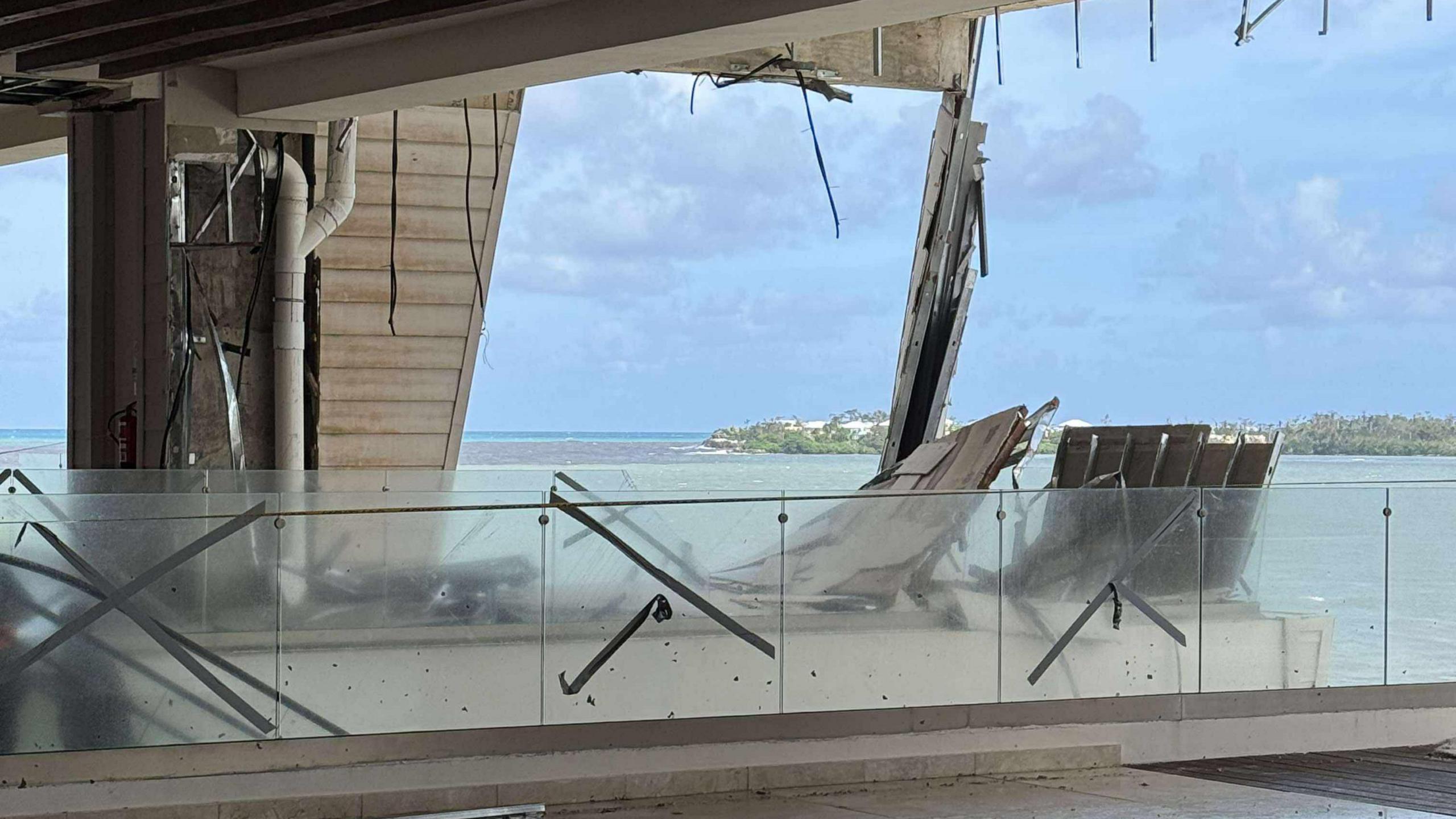 A photo taken from inside the hotel - it appears to be a patio of some sort with glass fences. Parts of the walls have collapsed or been ripped away, with wires and cables left dangling down. Beyond the wrecked wall is a view of the light blue Caribbean sea, with blue skies and a grassy headland also in the distance.