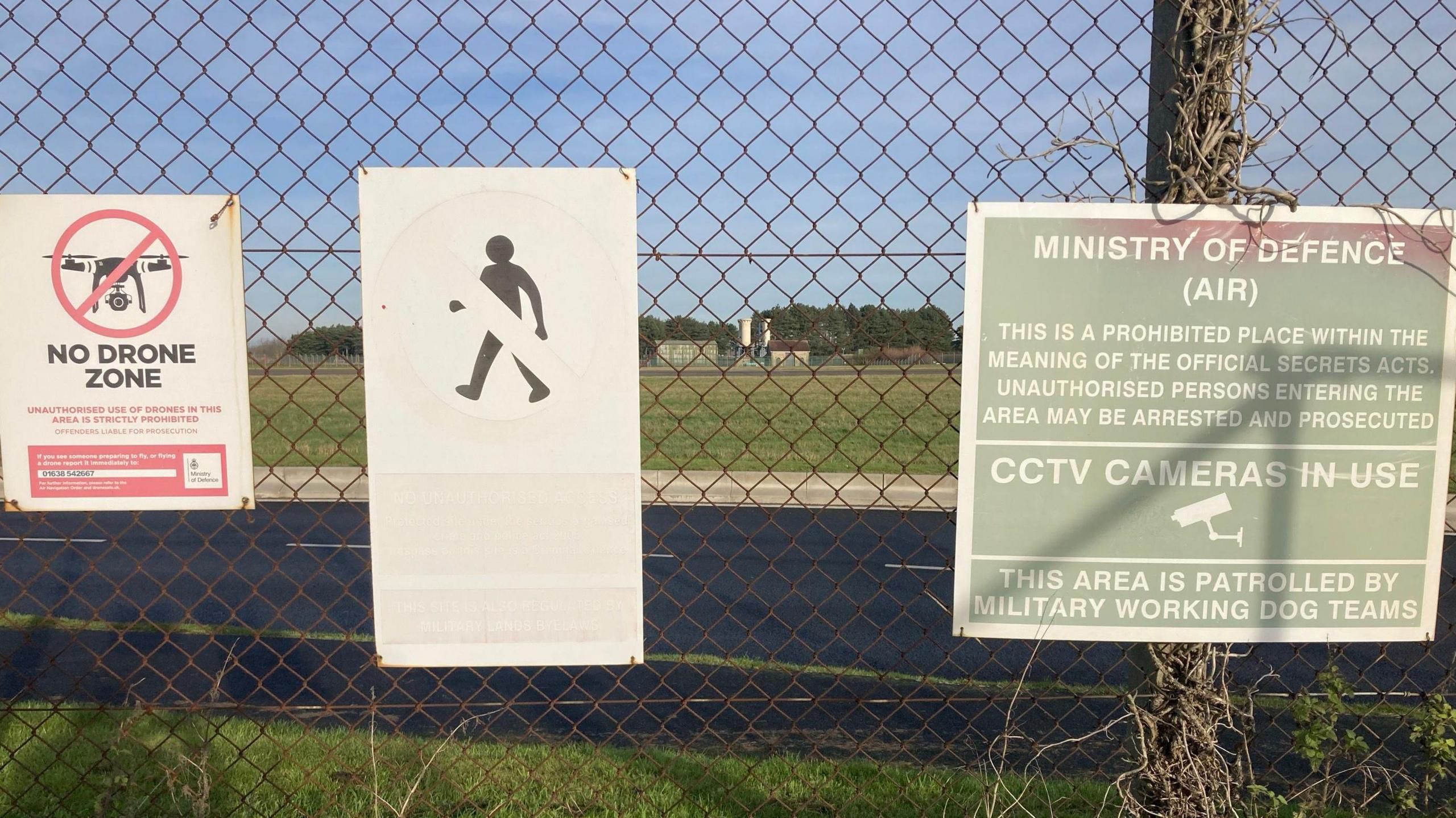 Three large signs sit on a fence warning people that the area is a no drone zone and there is a prohibited space behind the fence. An airbase sits behind the fence and several small buildings are seen in the distance. 