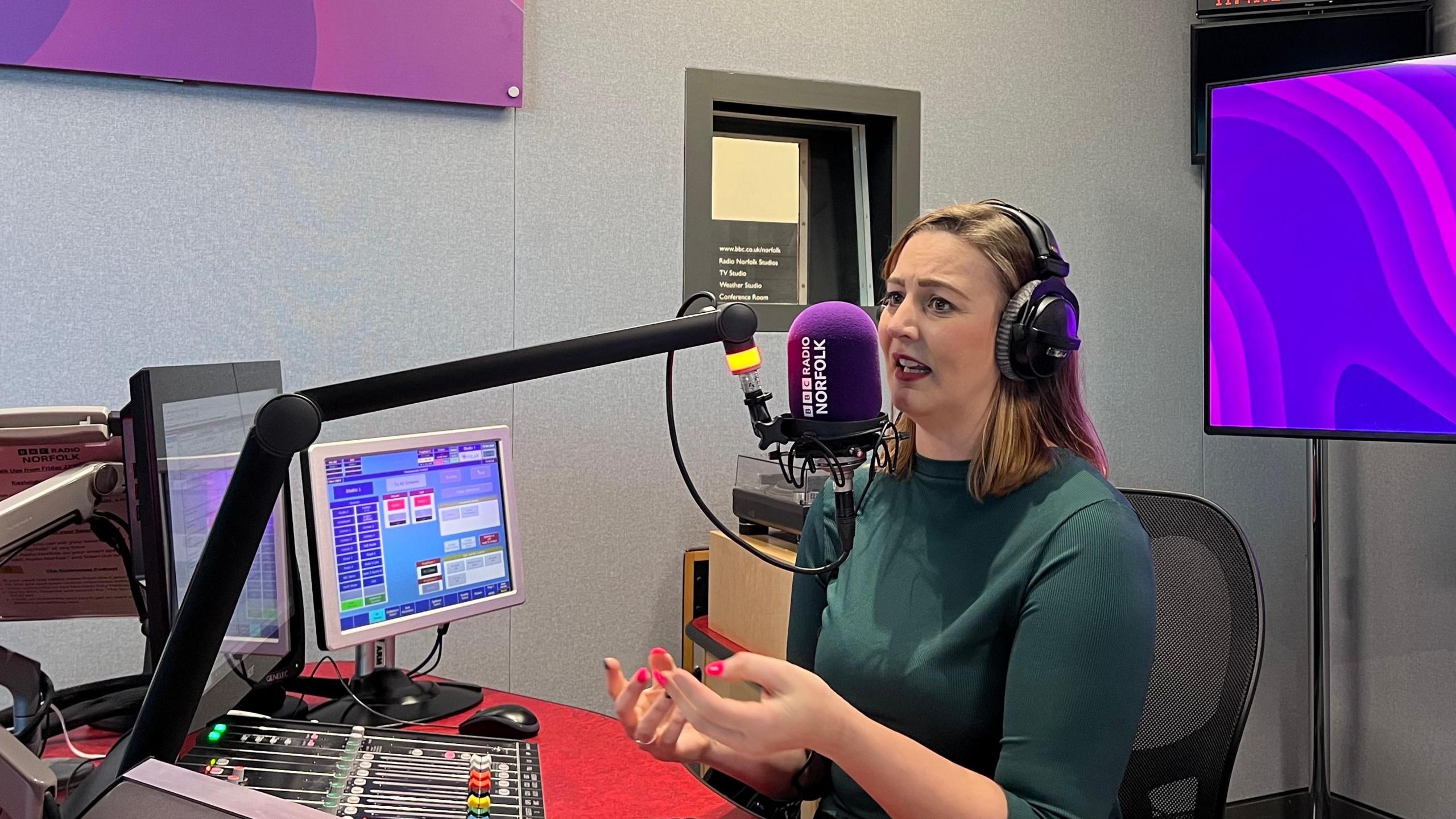 BBC Norfolk presenter Kayleigh Poacher sits in a radio studio asking questions. She is wearing a green top and large headphones as she sits talking into a microphone.