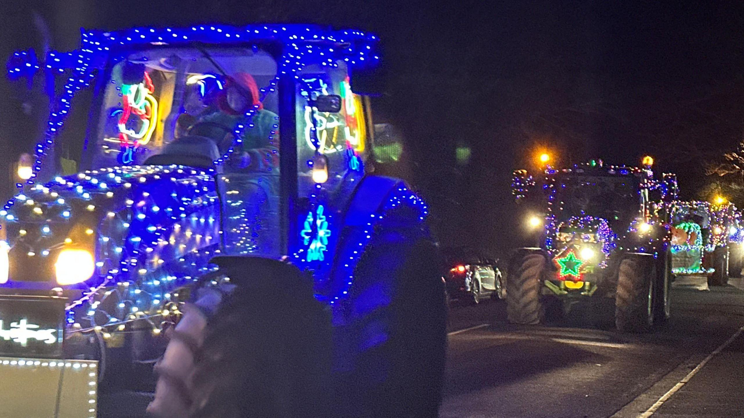 A tractor covered in blue lights in the fore, with others following behind, one has a red and green start on the front.