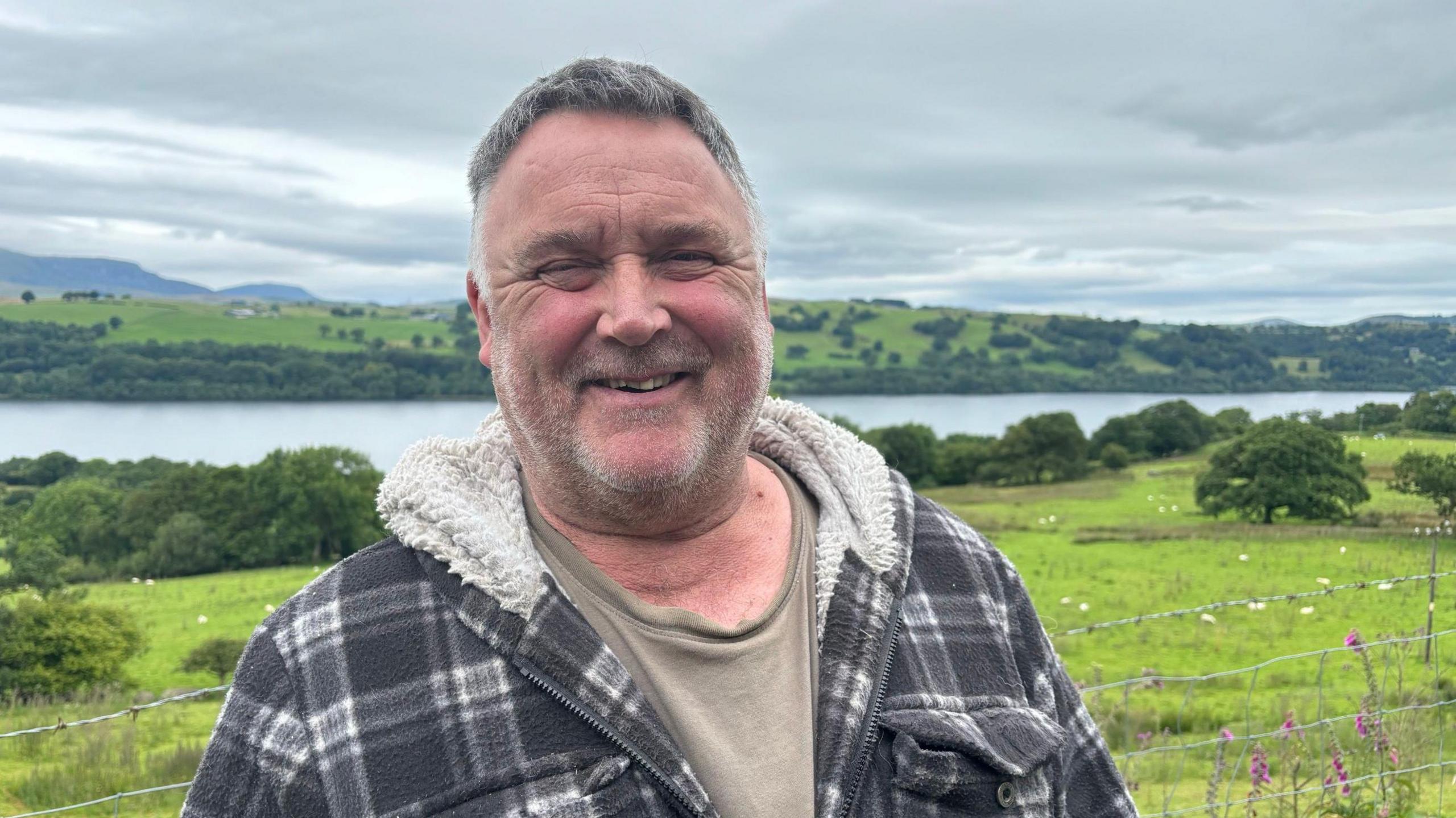 Geraint Roberts stands on farming land in front of a lake smiling at the camera