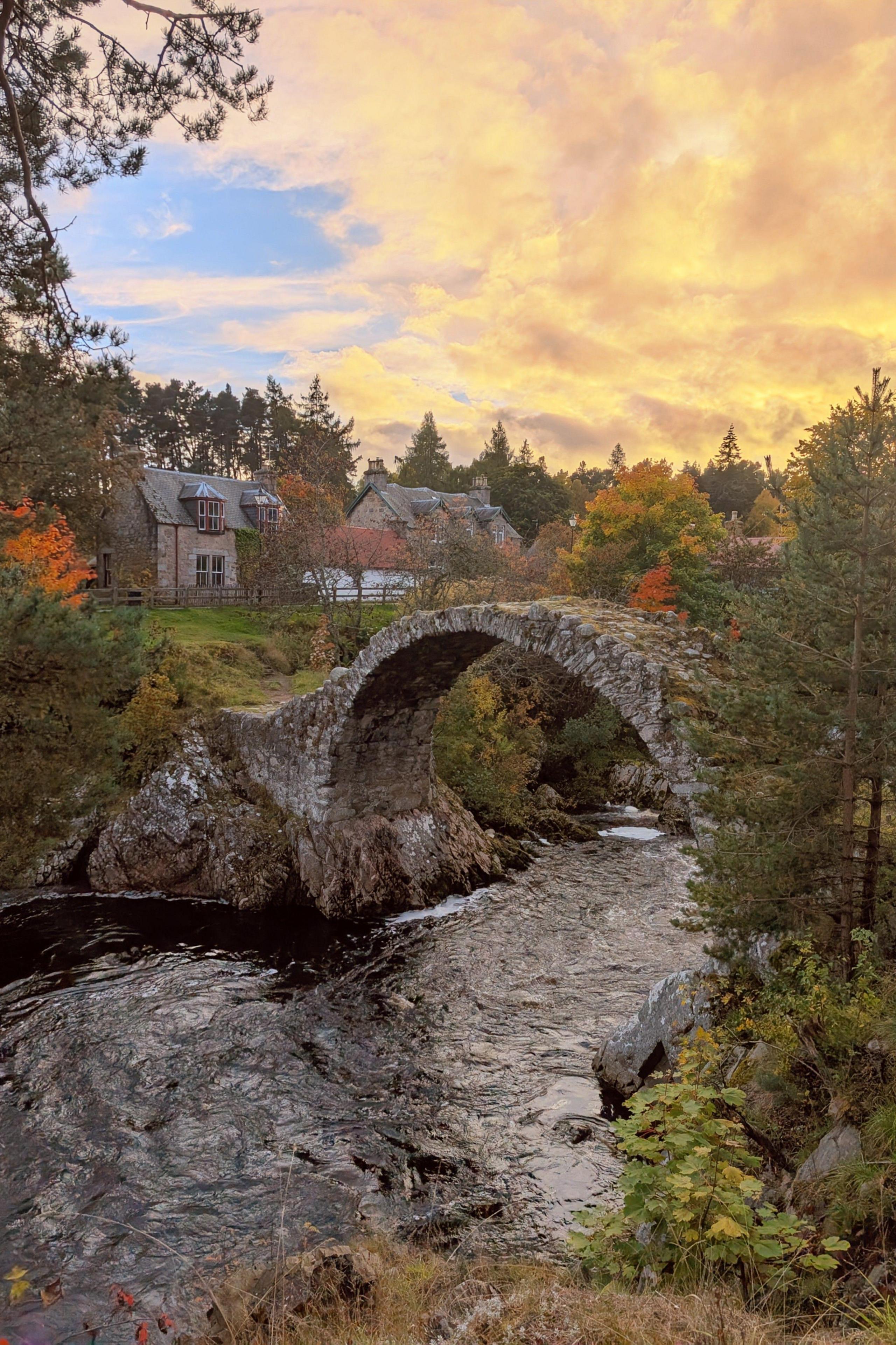 The sky is yellow and hangs over a stone arched bridge which has a river running under it.