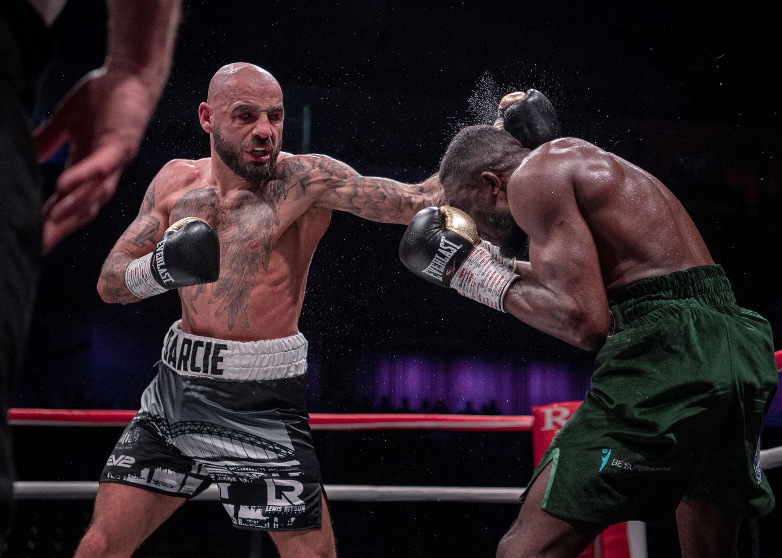 Bald, bearded boxer with chest and arm tattoos, and fierce face, lands a left-handed punch on his opponent's head.