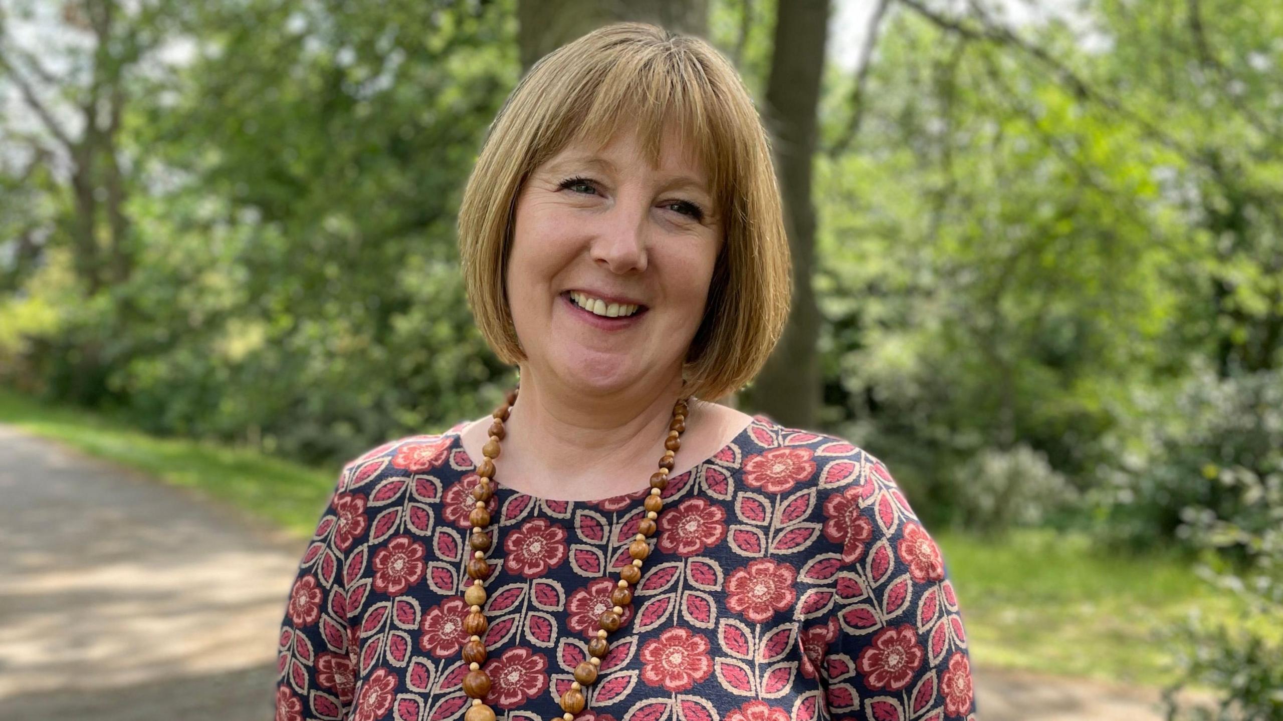 Charlotte Killeya, who has stage four melanoma, wearing a dress with red flowers on. She is outside at the Botanical Gardens in Sheffield and is smiling at the camera.