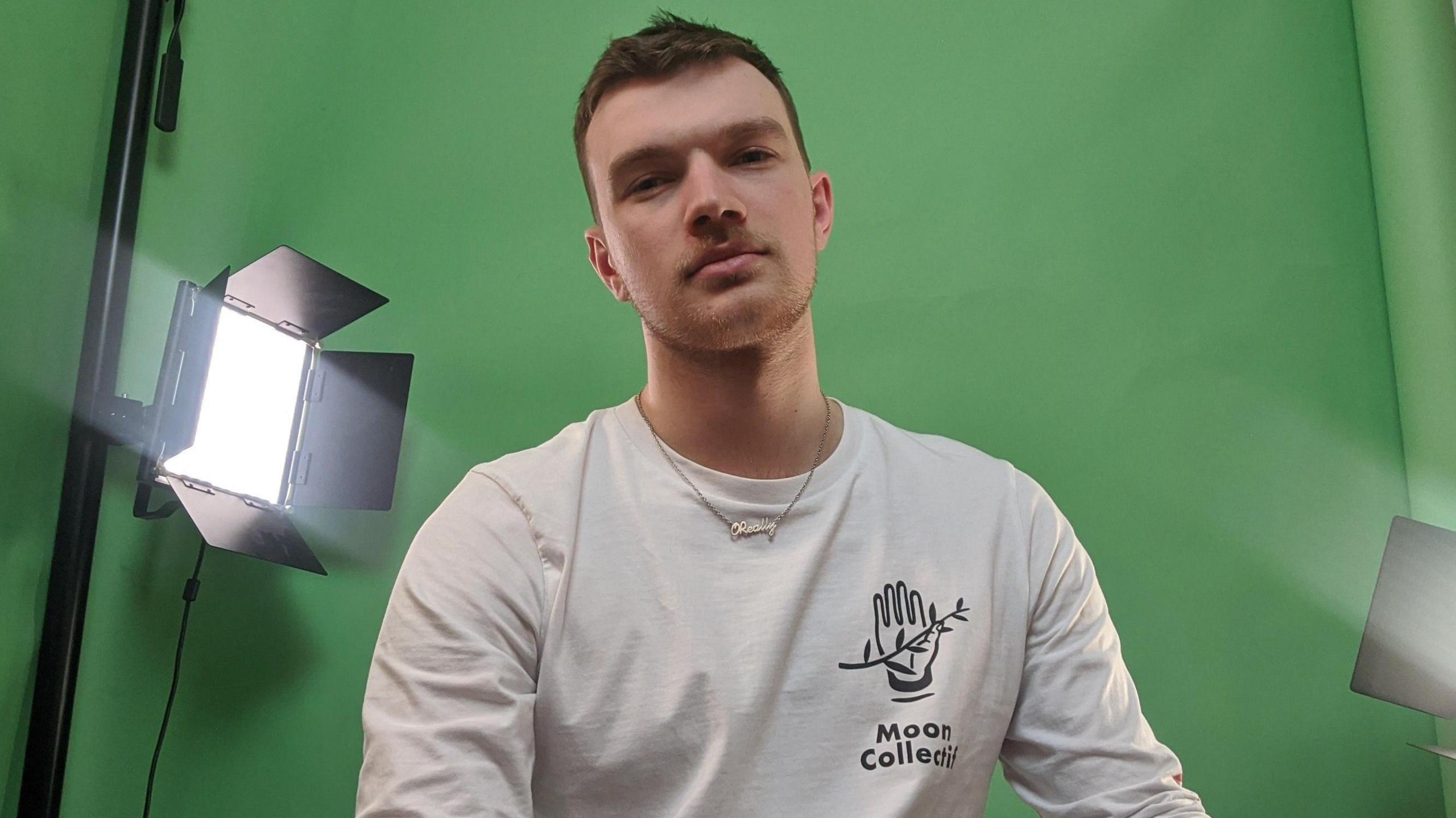 John O'Reilly looking at the camera with a green screen behind in the background, wearing a white top and necklace.