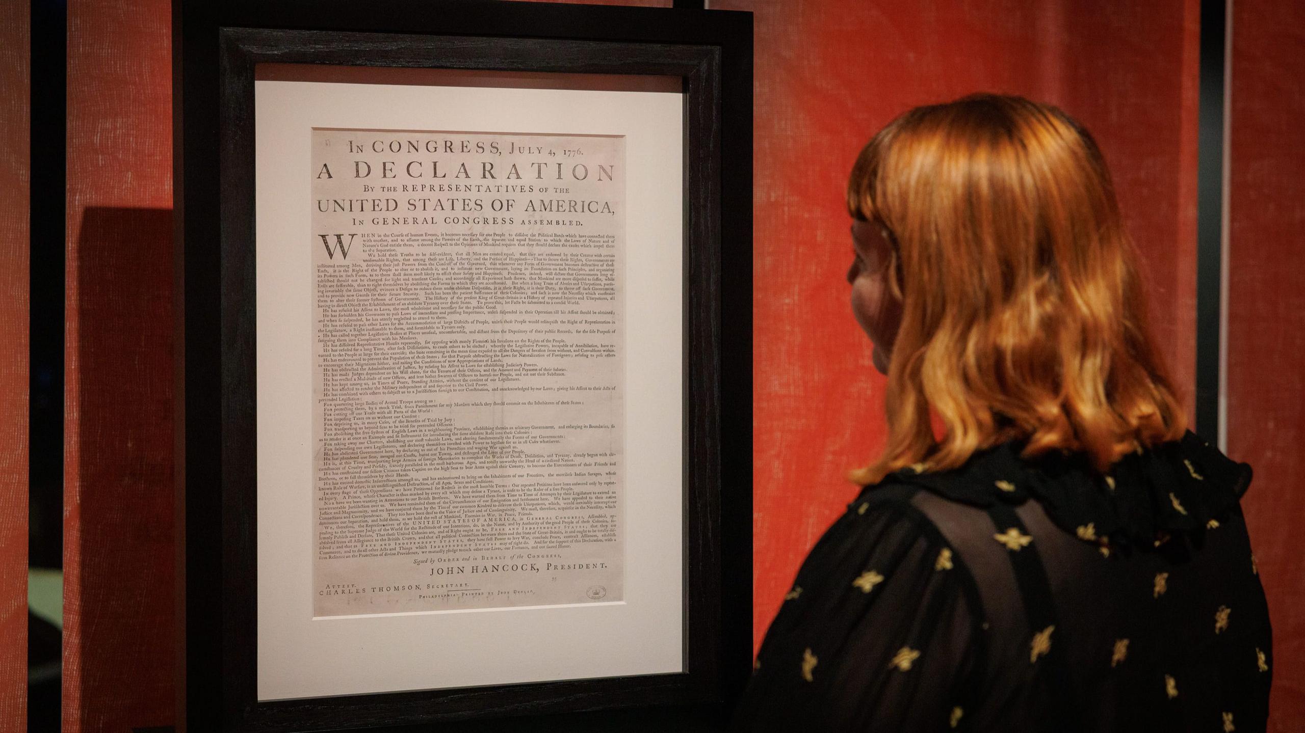 A woman with red hair looks at a framed copy of the Declaration of Independence in a brightly-lit room.