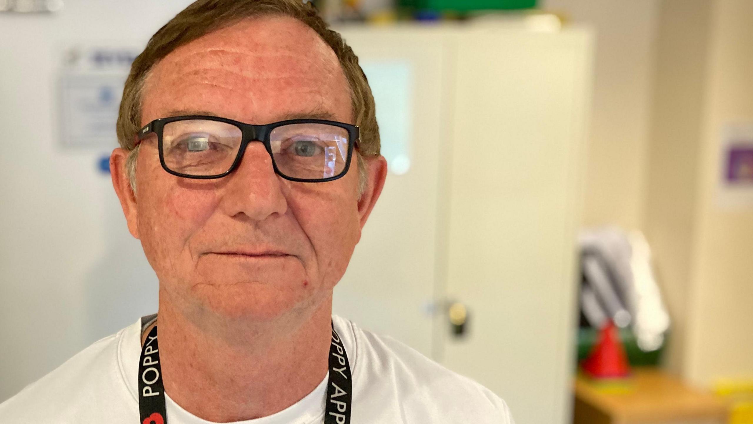Steve Foley looks straight at the camera and smiles. He has short brown hair, glasses, a black lanyard around his neck and he wears a white T-shirt. 