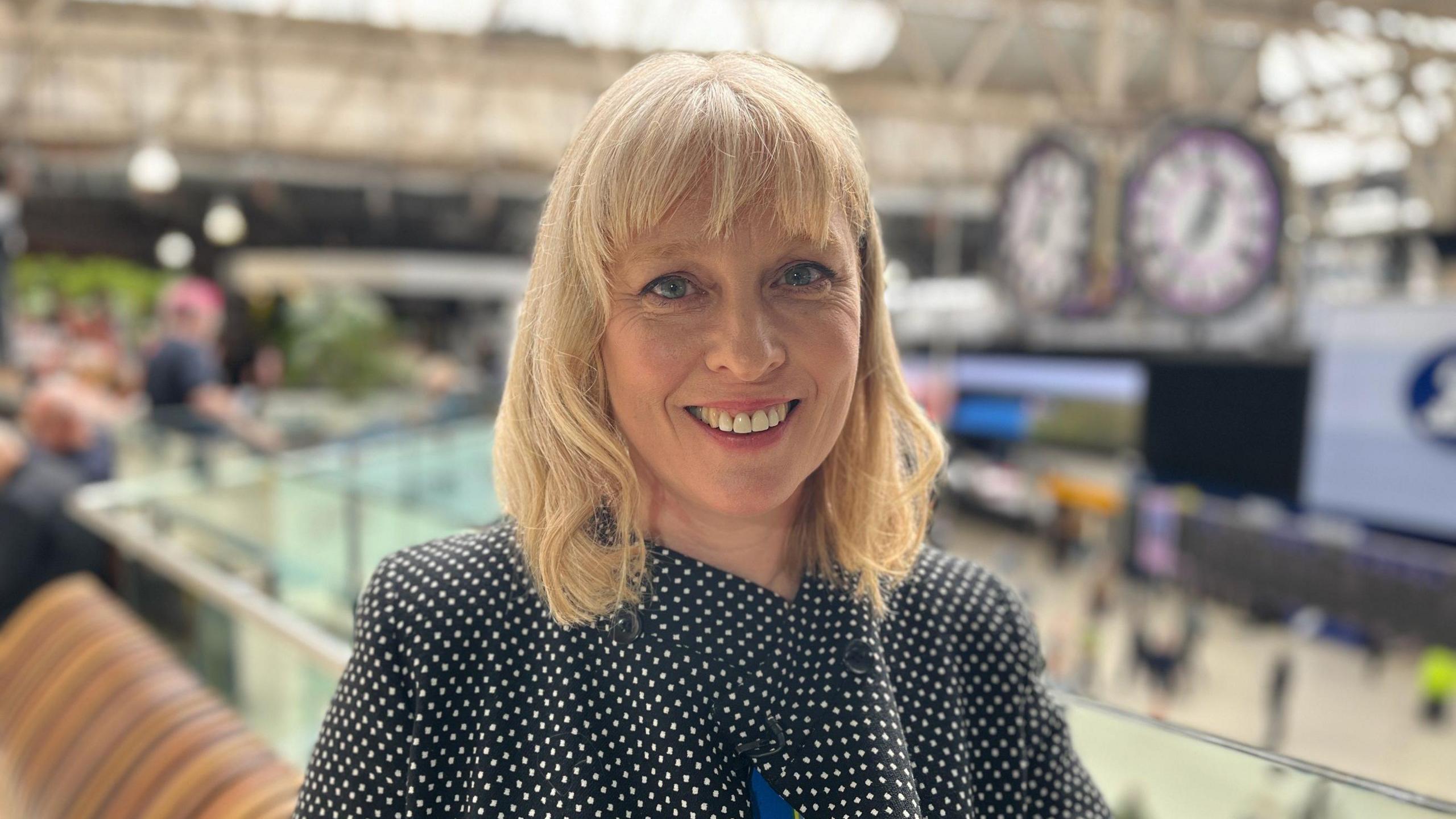 Sophie Woolley is standing in a busy train station, she is upstairs with a view of the lower platform area below and large clocks suspended from the ceiling. Behind her are also passengers walking. Sophie has mid length blonde hair, wears a pink lipstick and a black blouse with white polka dots