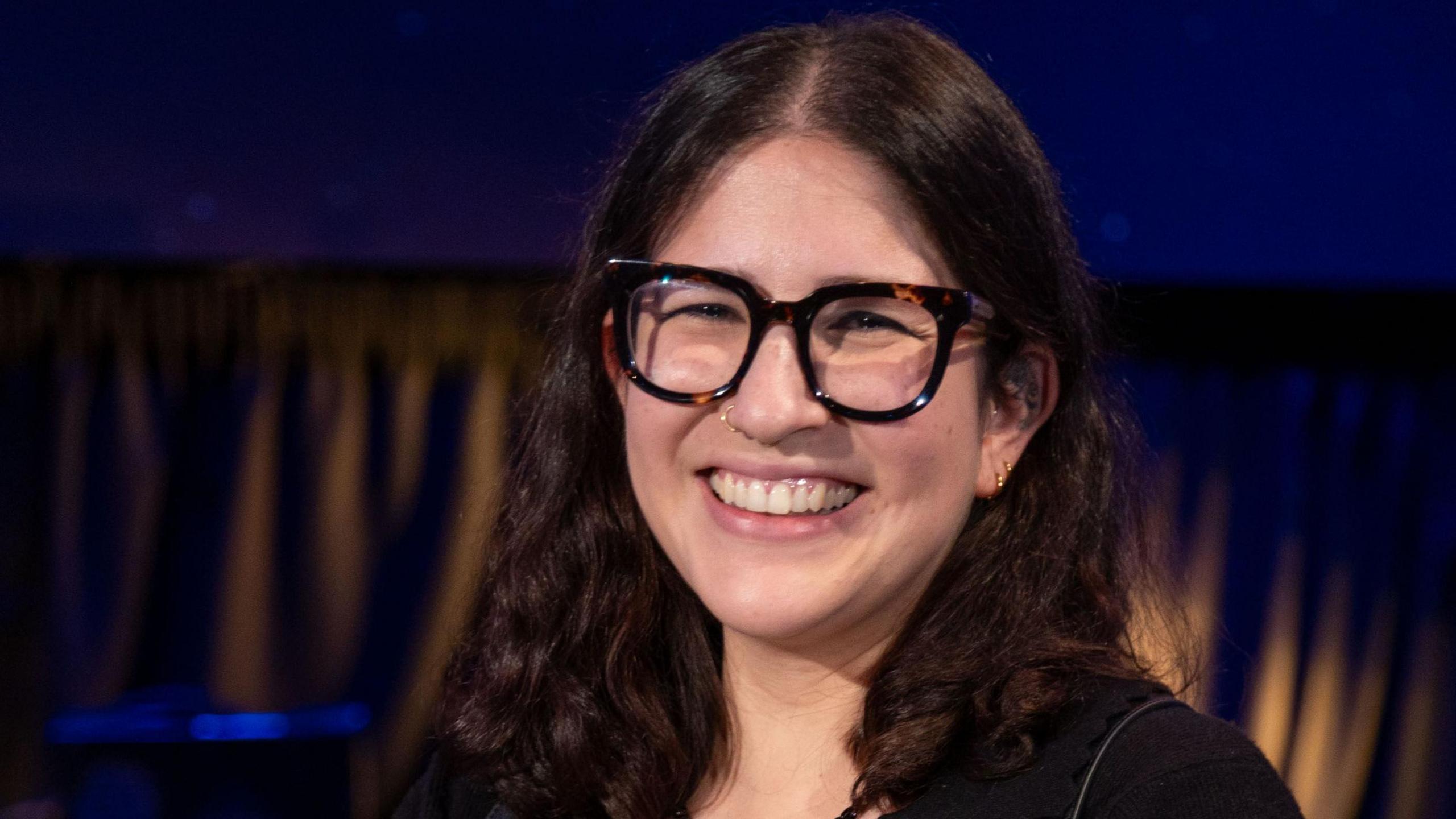 A woman wearing thick-rimmed dark glasses with long curly hair smiles. 