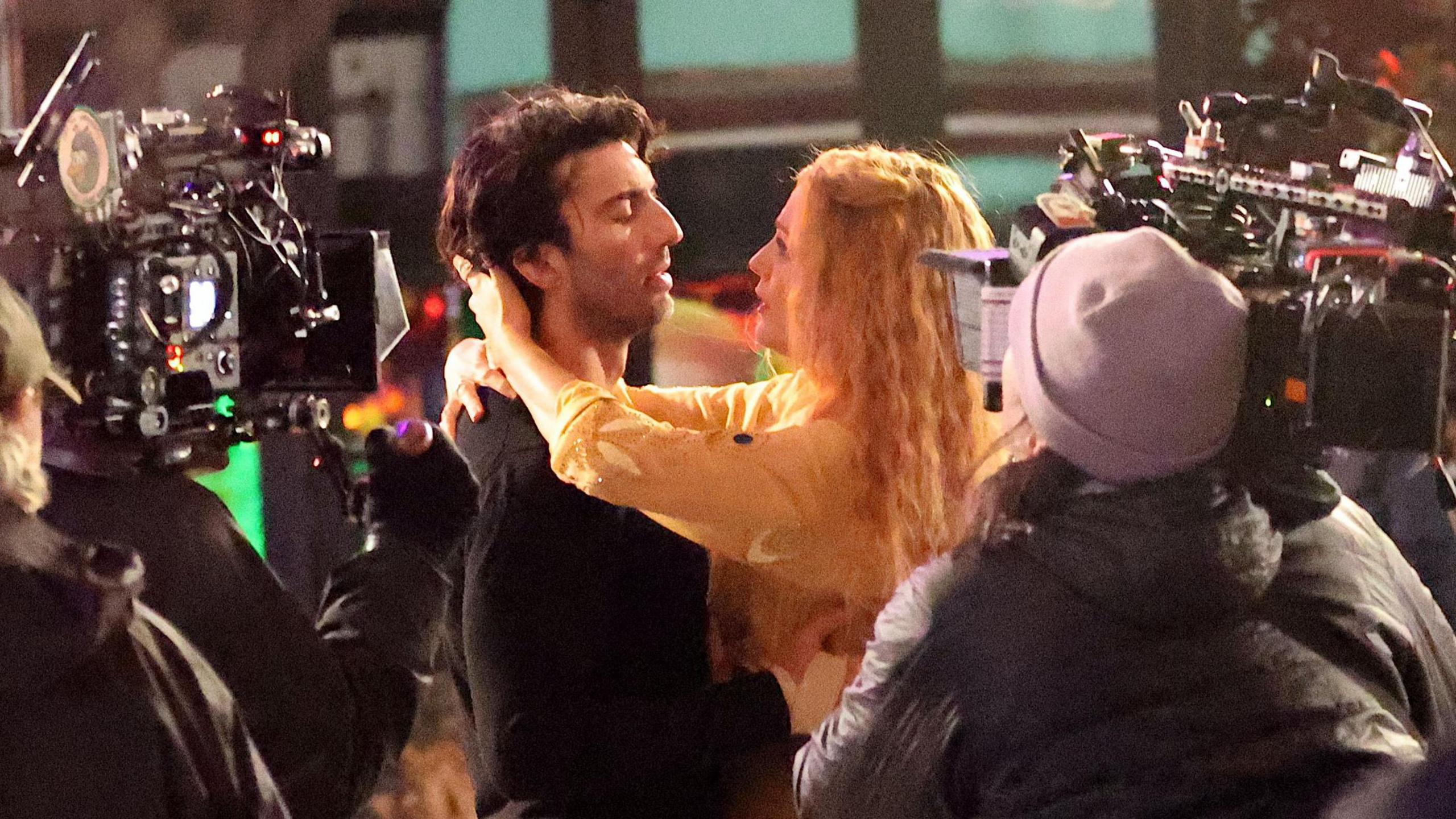 Justin Baldoni and Blake Lively pictured on set for It Ends With Us. Justin has floppy brown hair and wears a black jumper. Blake, who has crimped blonde hair, has her arms around his neck looking into his eyes. They're surrounded by camera operators filming them. 