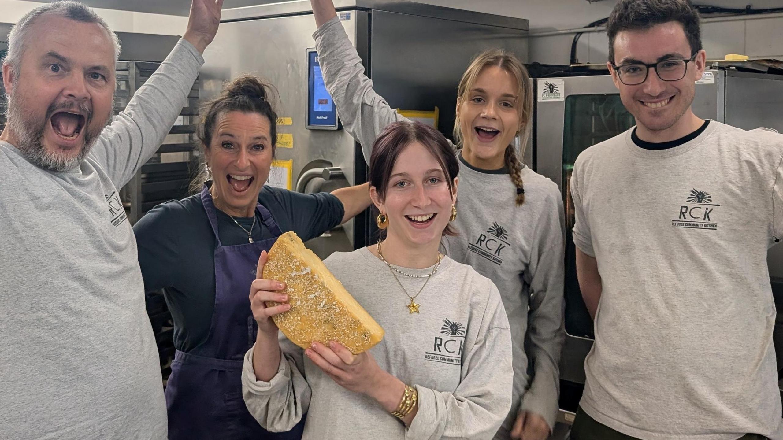 Image of a group of people cheering in a commercial kitchen, with central female figure holding a round wheel of cheese.