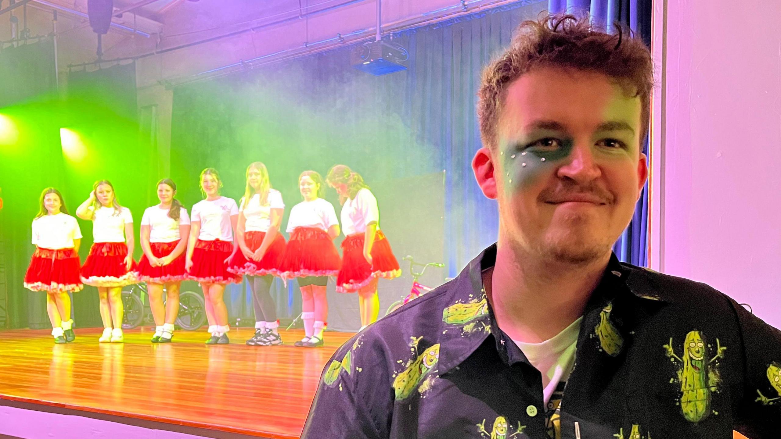 Jordan Winn stands in the foreground with a line of dancers in identical white tops and red skirts in the background. Jordan is wearing green make up on his eyes and a black shirt.