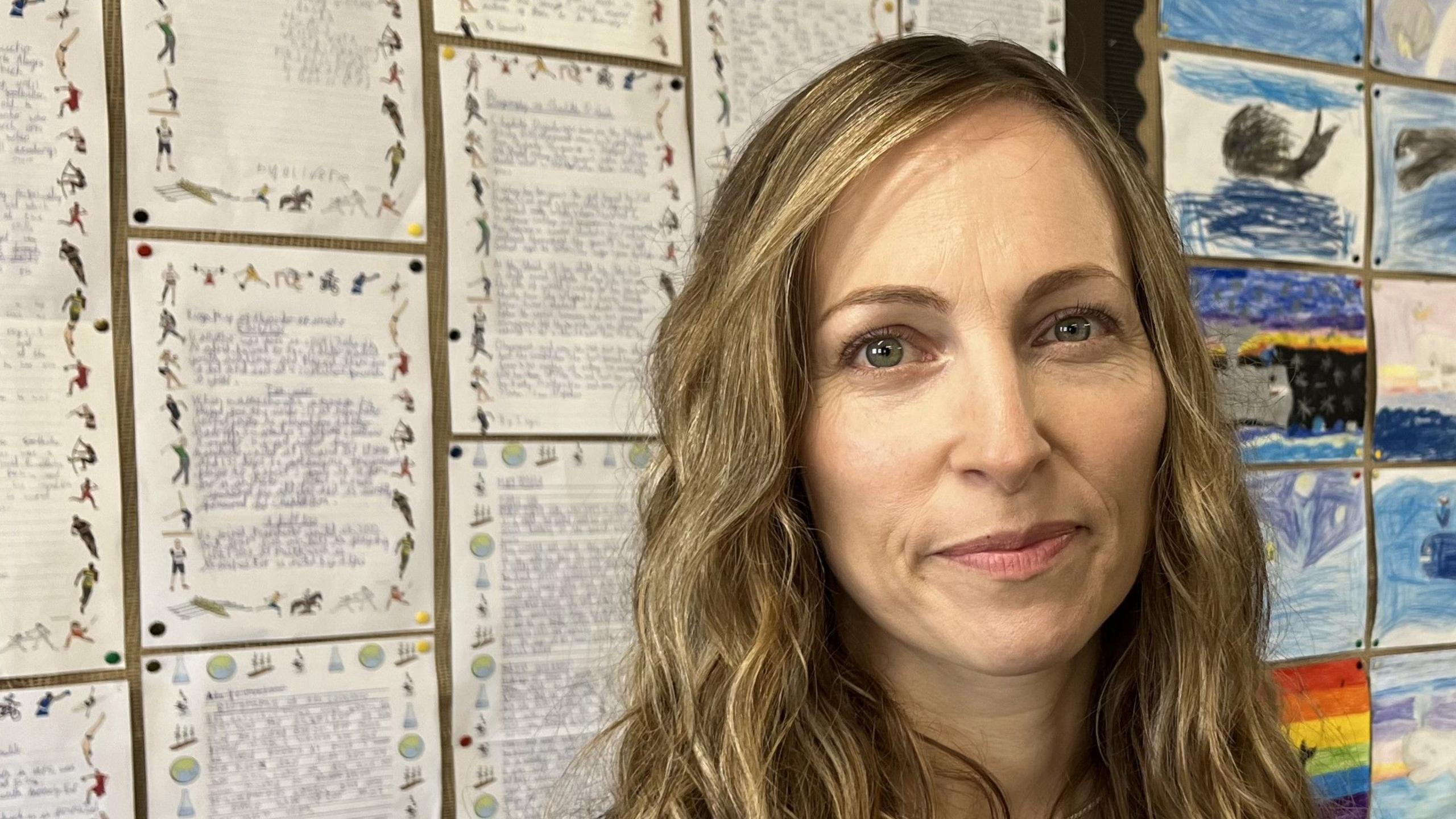 Jules Stops, who has long blonde hair, stands in front of a noticeboard with children's writing and drawing pinned to it.