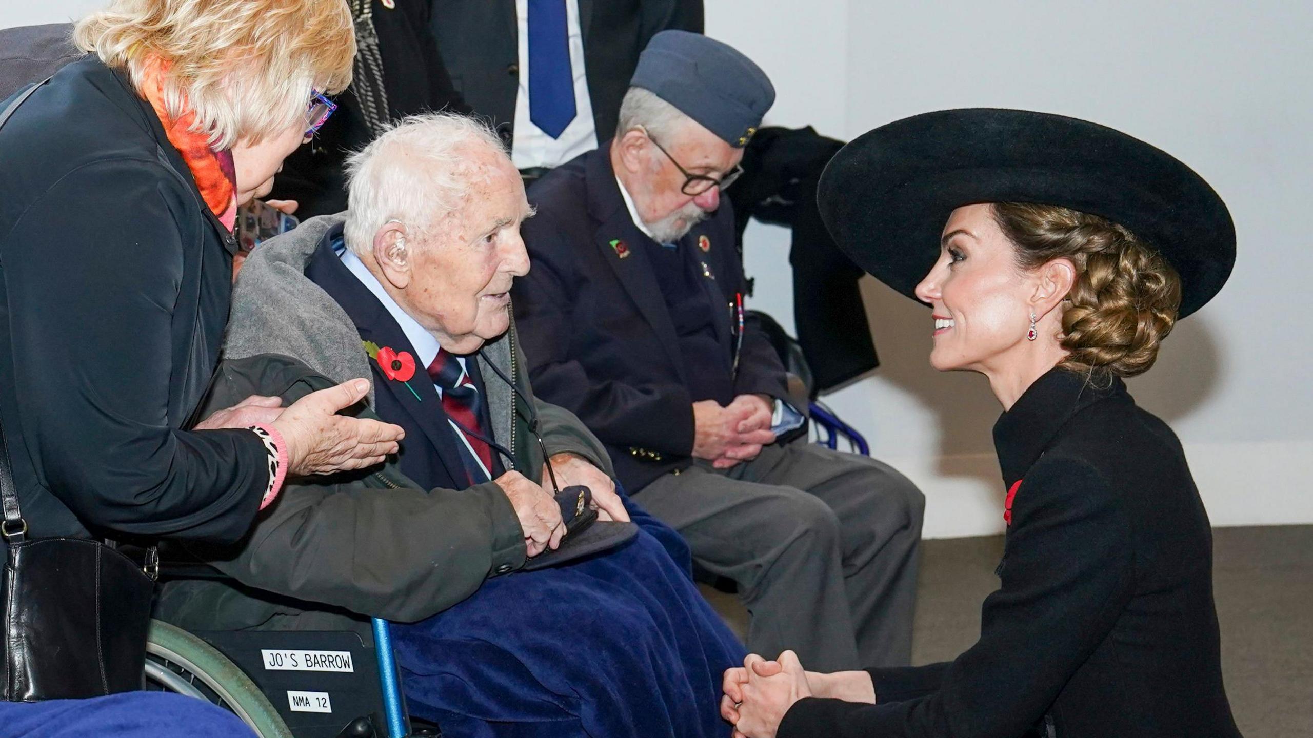 An elderly man in a wheelchair is speaking to the Princess of Wales. She is dressed in a large black hat, long black coat with a red poppy and her brown hair is tied up. She is kneeling to speak to the man, who has short white hair and is dressed in a navy suit, also wearing a poppy and has a blue blanket covering his legs. He is sat next to anothe man in a wheelchair wearing grey trousers, a navy suit and a blue beree. There is also a woman with blonde, short hair, a dark coat and orange scarf leaning over speaking to the princess. They are in a room with white walls and grey carpet.