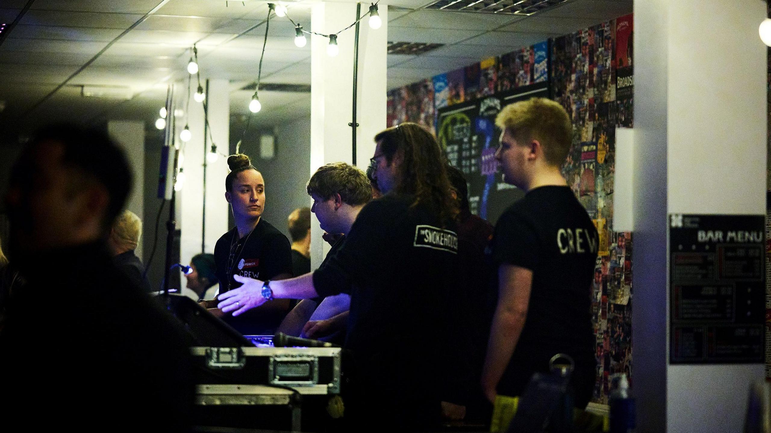 A small group of people in black 'crew' T-shirts stand around a mixing desk in a music venue. There are lights with exposed bulbs hanging in the background. 