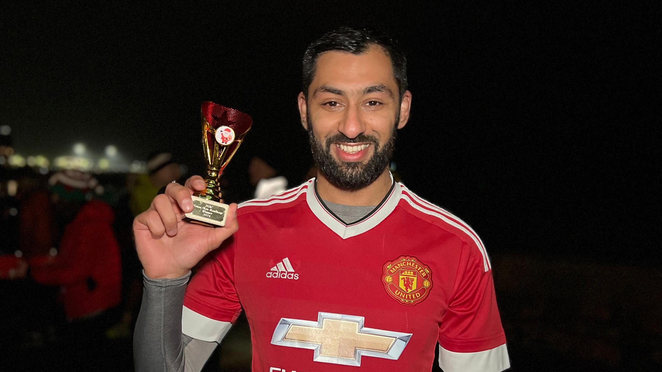 Ismail smiles at the camera as he holds the trophy after the race. He's wearing a Manchester United football top with a Jersey Spartan AC race number on it. He as a grey lycra top under his shirt. He has a short dark beard and short dark hair. Runners are standing behind him in the dark.