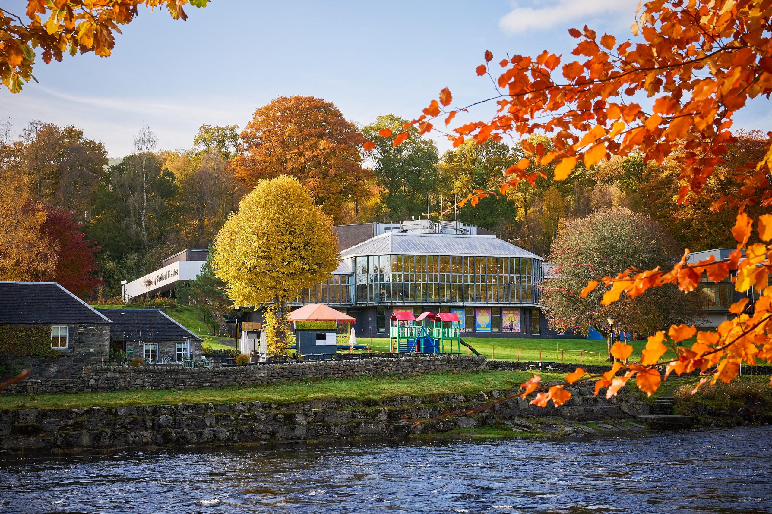 Pitlochry Festival Theatre building as seen from river. It is autumn and the leaves are red and orange