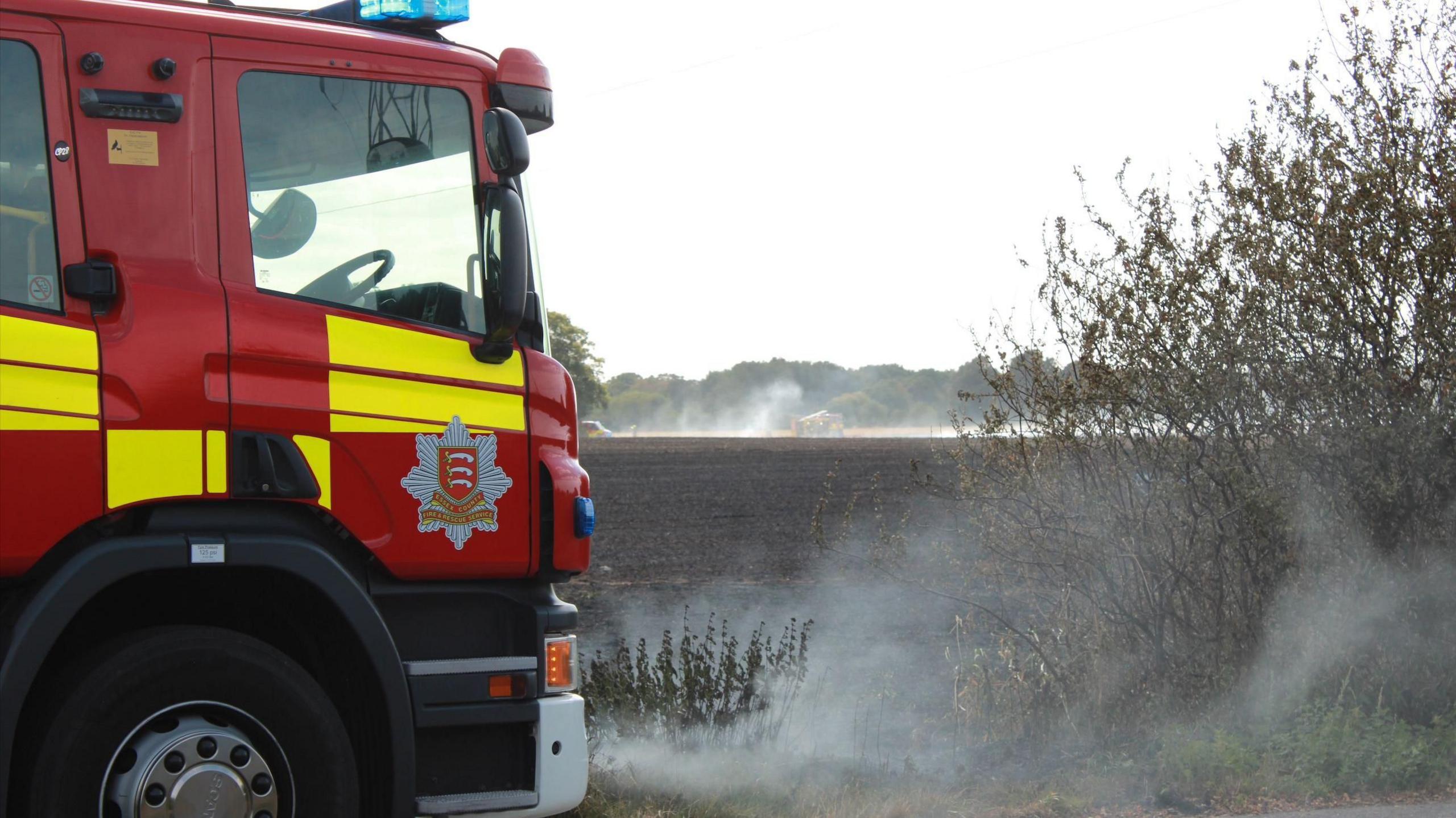 A red and yellow fire engine with fields behind it and smoke billowing up