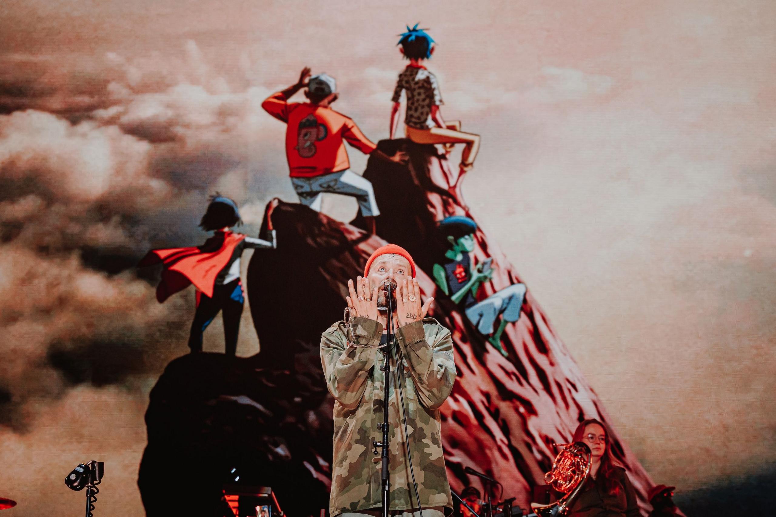 Damon Albarn sings with his hands raised to his face. In the background, a video screen shows an image of Gorillaz band members standing at the peak of a mountain.