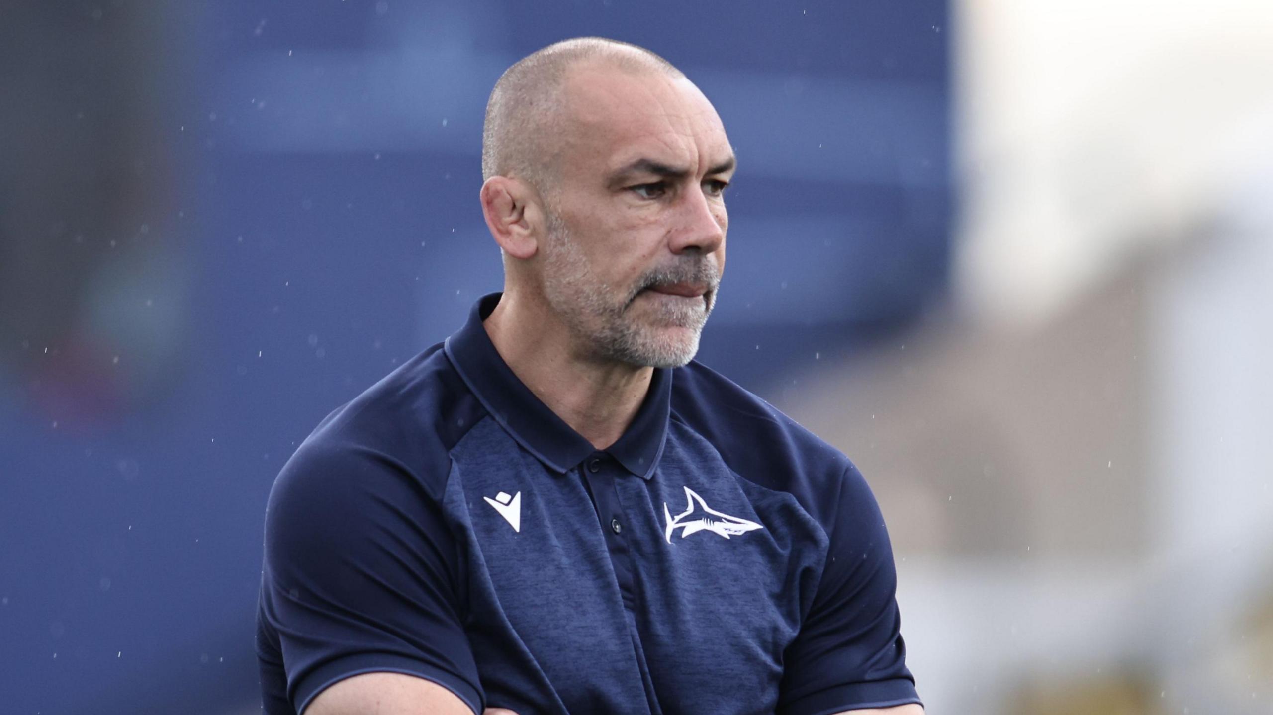 Alex Sanderson looking pensive, wearing a navy blue Sale Sharks t-shirt and stood with his arms folded