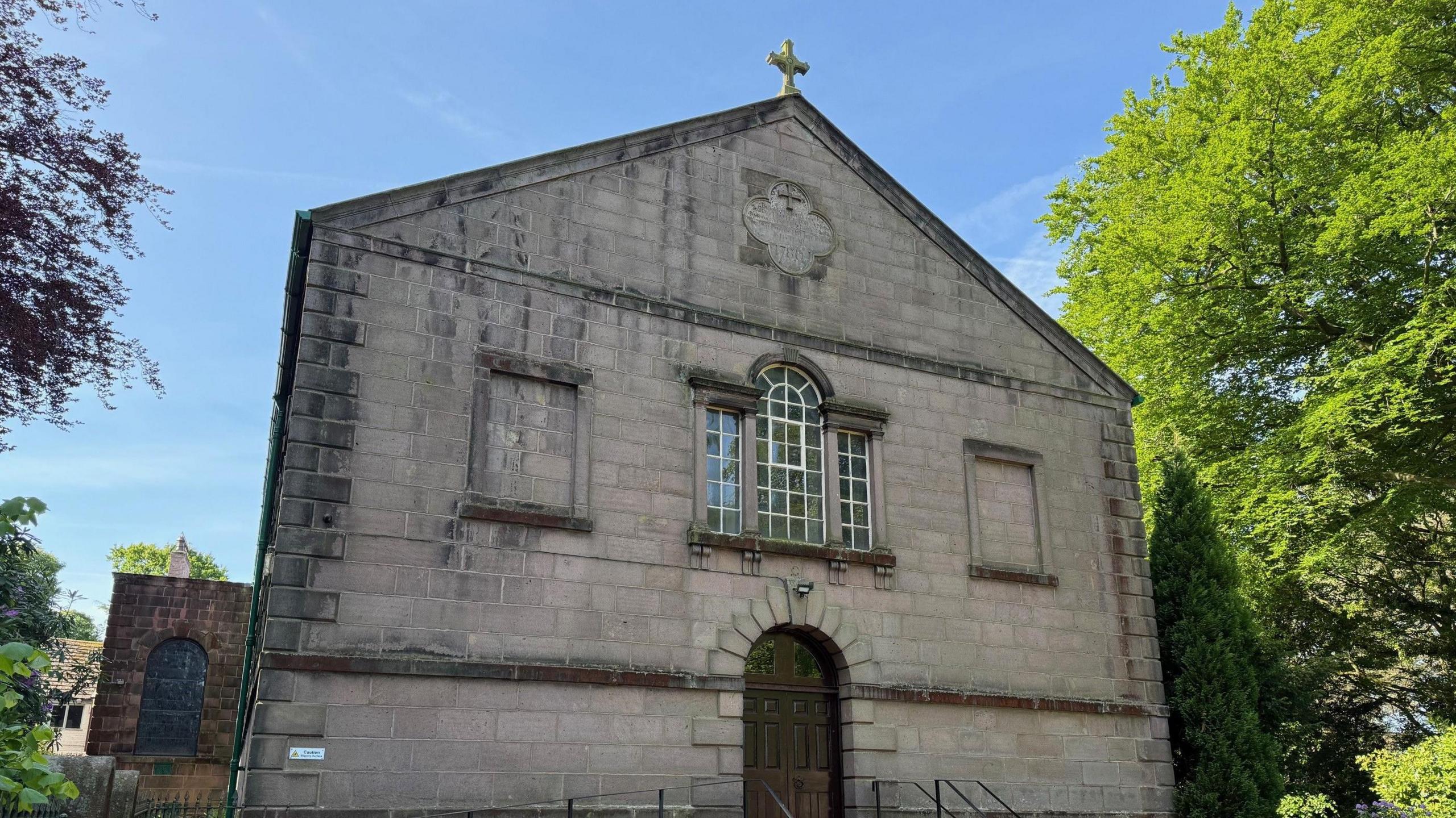 An old grey brick church is surrounded by large green trees.