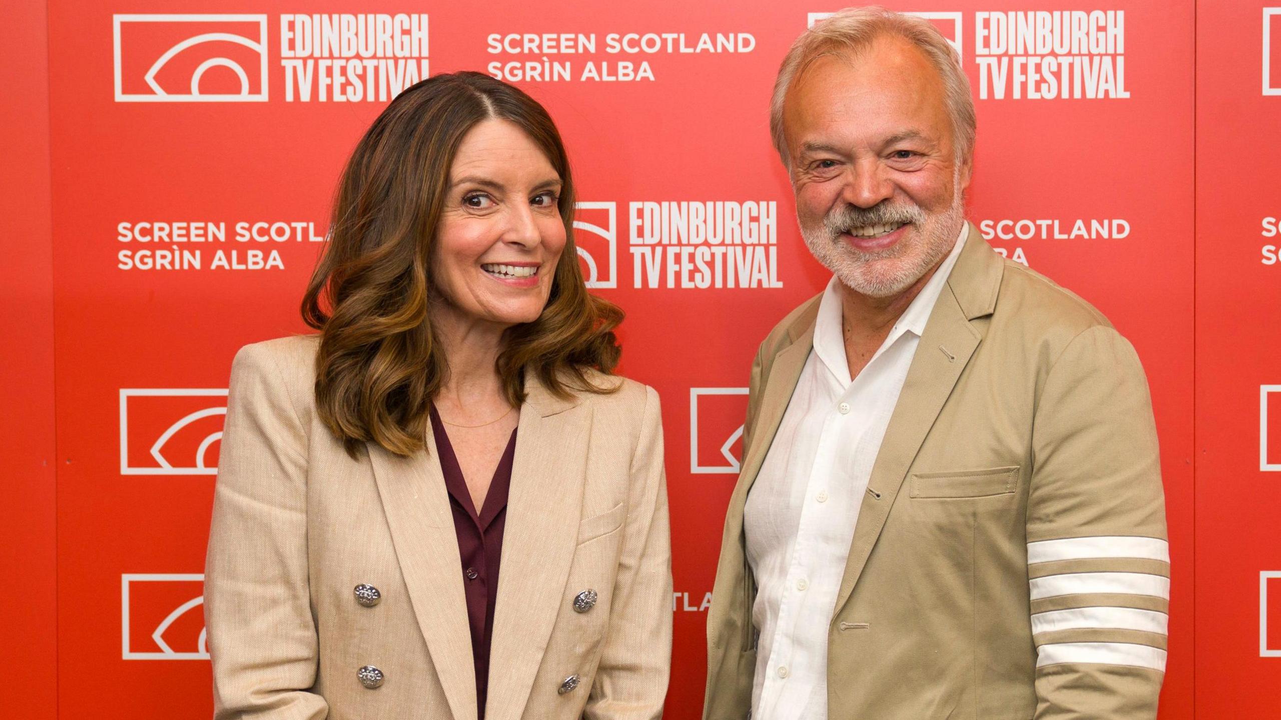 Tina Fey, who has log brown hair and is wearing a maroon blouse with a beige blazer, standing against a bright red backdrop multiple white logos including "Edinburgh TV Festival" and "Screen Scotland". She is smiling. Graham Norton, who has grey hair and a beard is standing grinning next to her. He is wearing a beige blazer with four white stripes on the left arm.