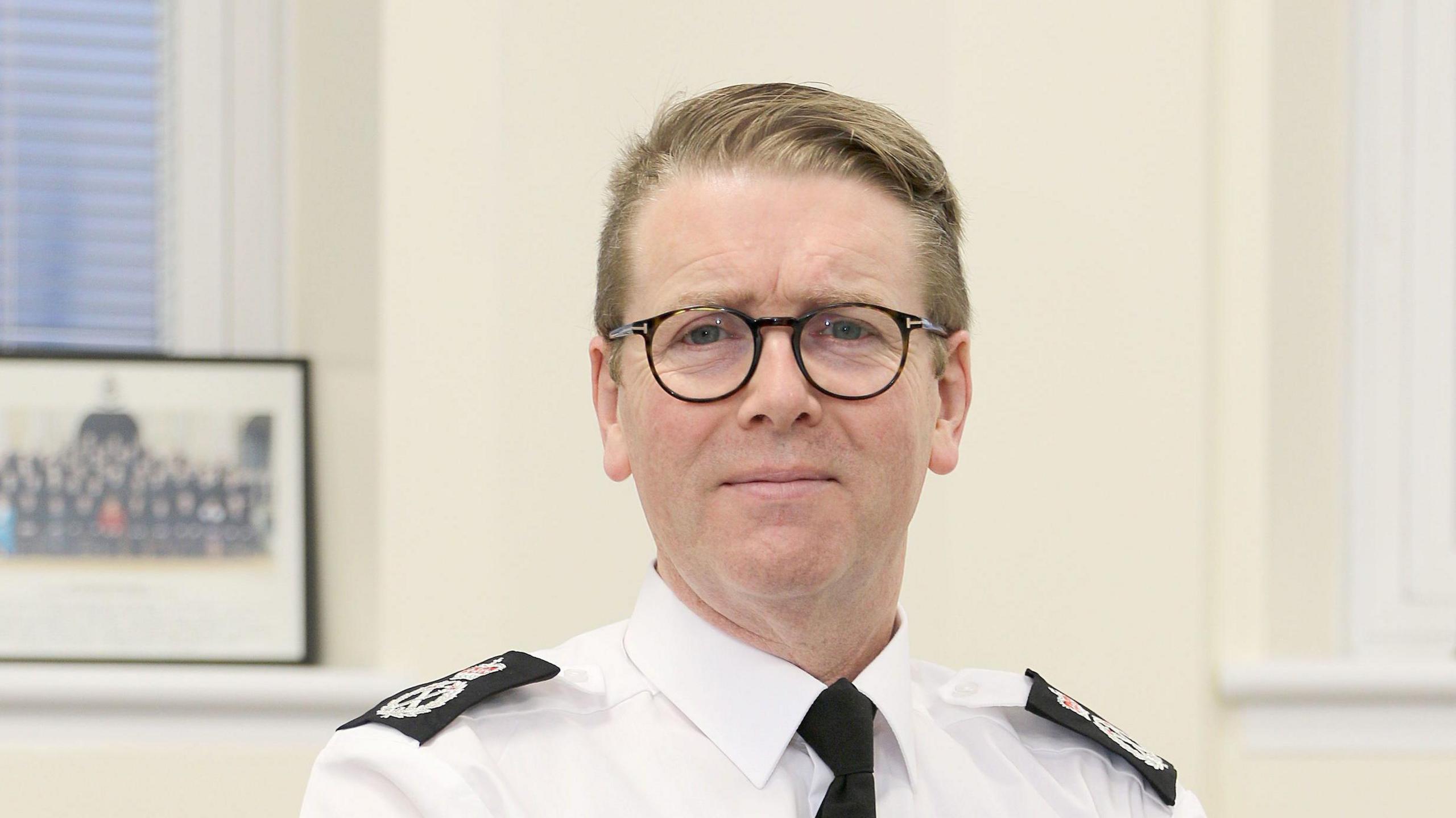 Will Kerr is pictured wearing a white shirt with a black tie and police shoulder numbers. The photograph only shows him from the shoulders up and he is looking directly at the camera. He appears to be in an office with cream walls. He is wearing dark round glasses and has short dark blond hair.