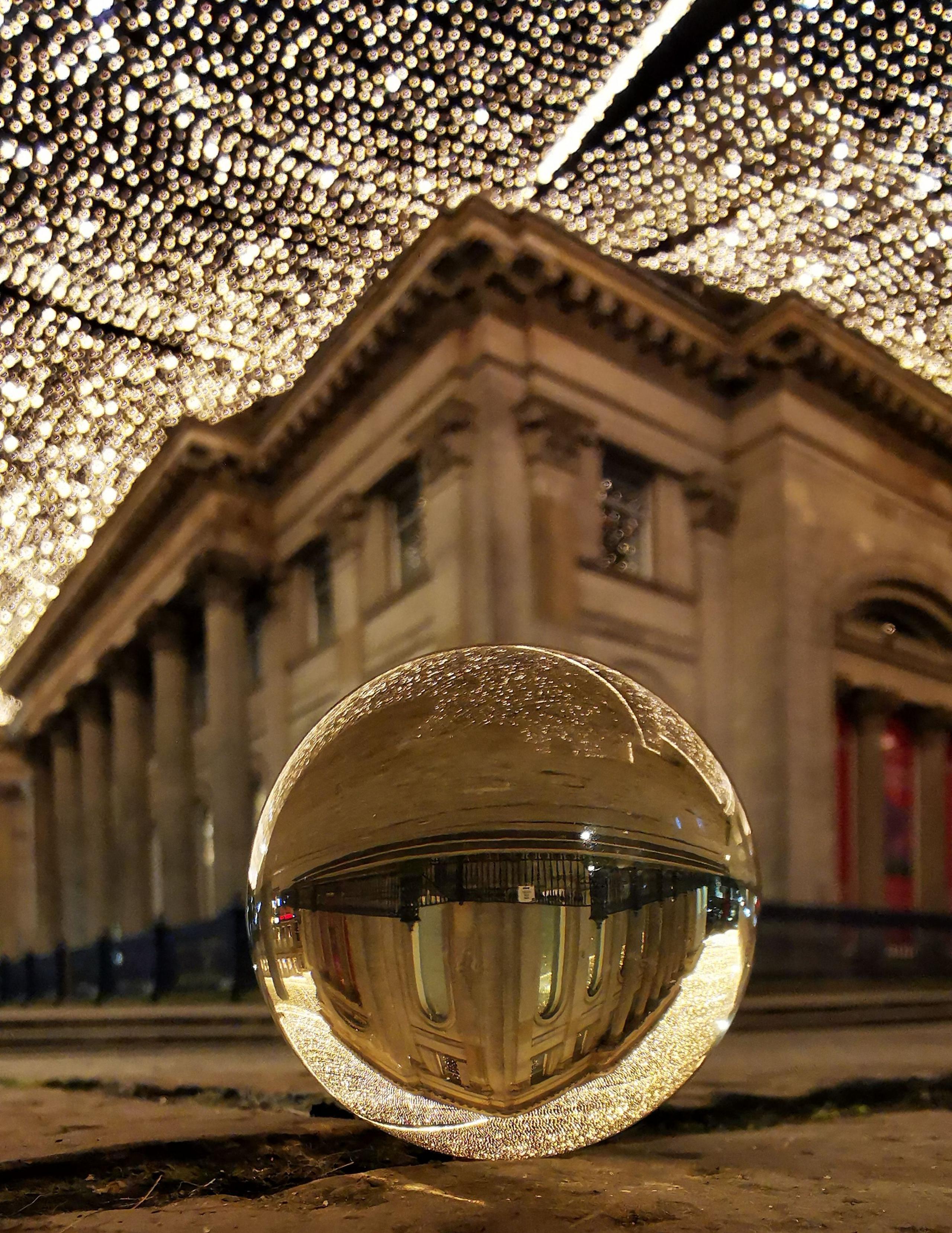 Evening image of the Gallery of Modern Art in Glasgow, taken from street level with sheets of fairy lights hung above Royal Exchange Square. A decorative glass ball is in the foreground and the museum is inverted in the reflection of the ball.