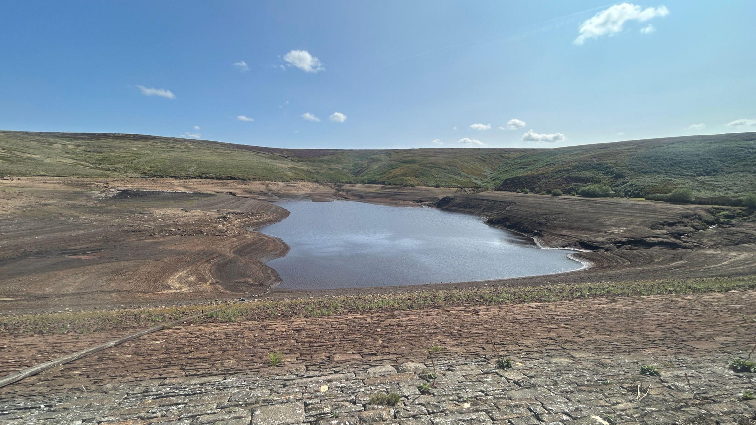 A reservoir in a rural location and water levels are very low. There is blue sky above and hills in the background.