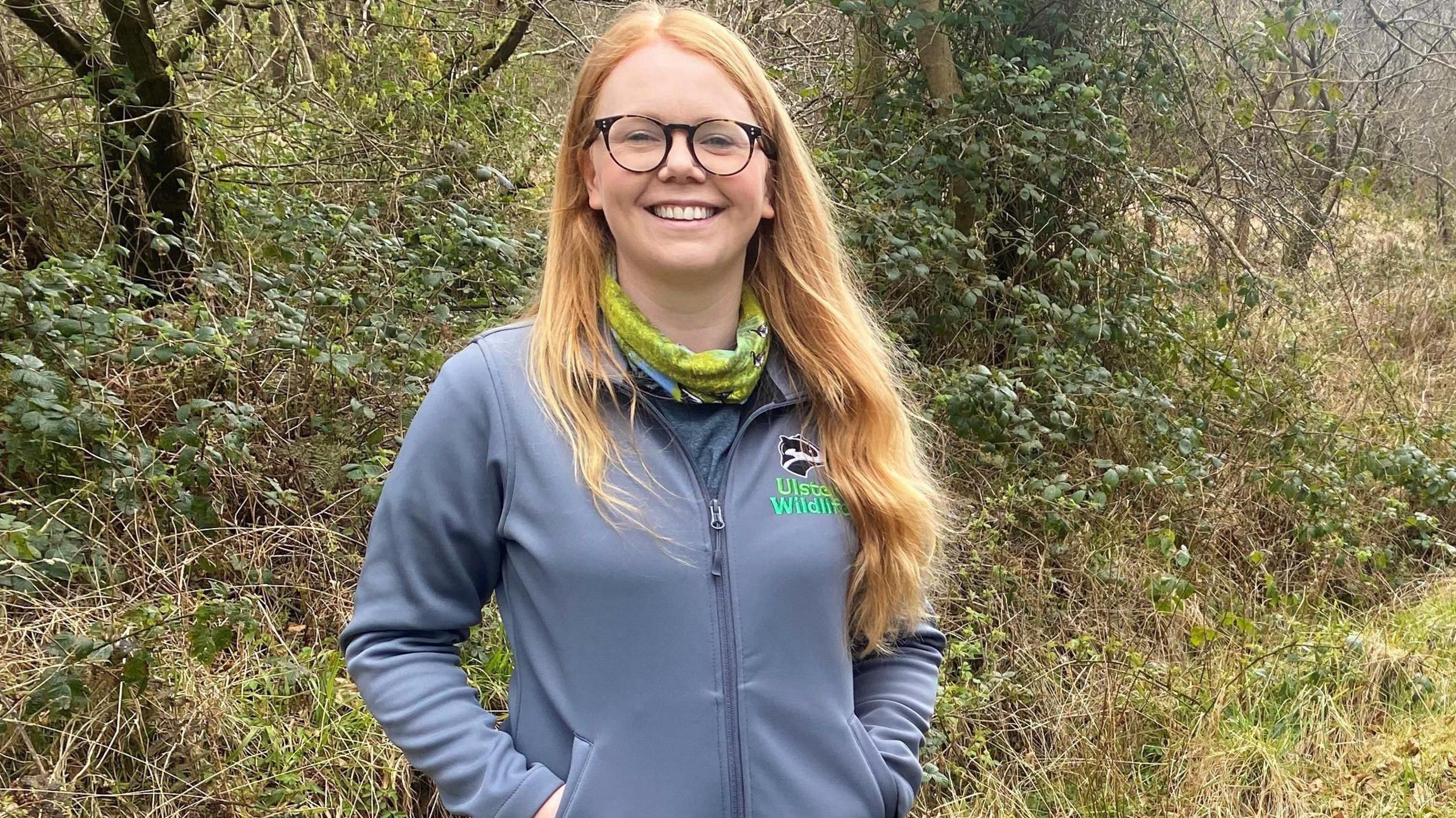 Katy is standing in a wooded area. She wears glasses and has long auburn hair. She's wearing an Ulster Wildlife jacket and a scarf.