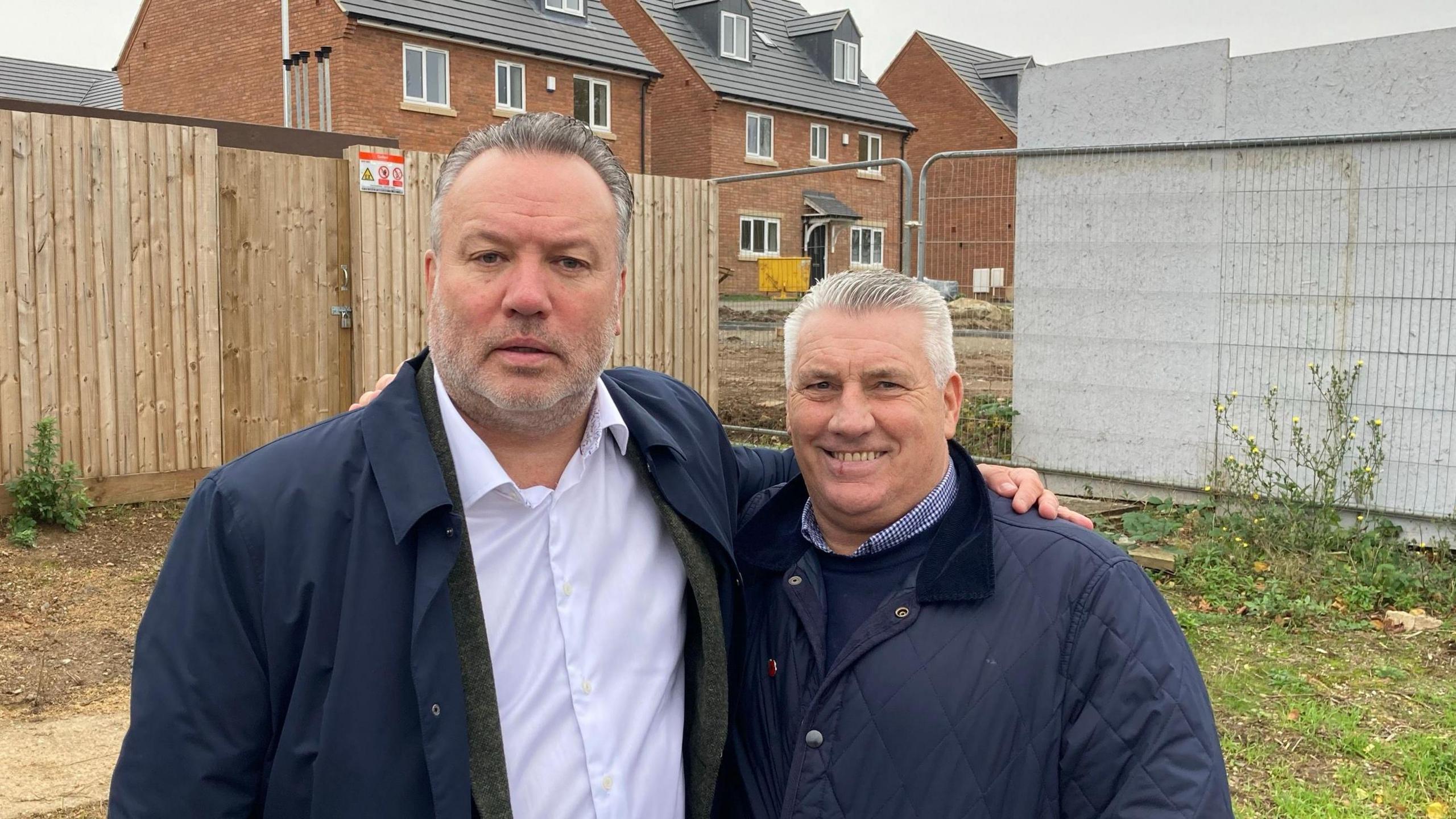 Kevin Shoemake and Dean Thomas standing side by side with their arms around each other's shoulders. Shoemake is wearing a navy trench coat which is unbuttoned and showing a white buttoned shirt underneath. Thomas is in a blue coat and smiling at the camera.