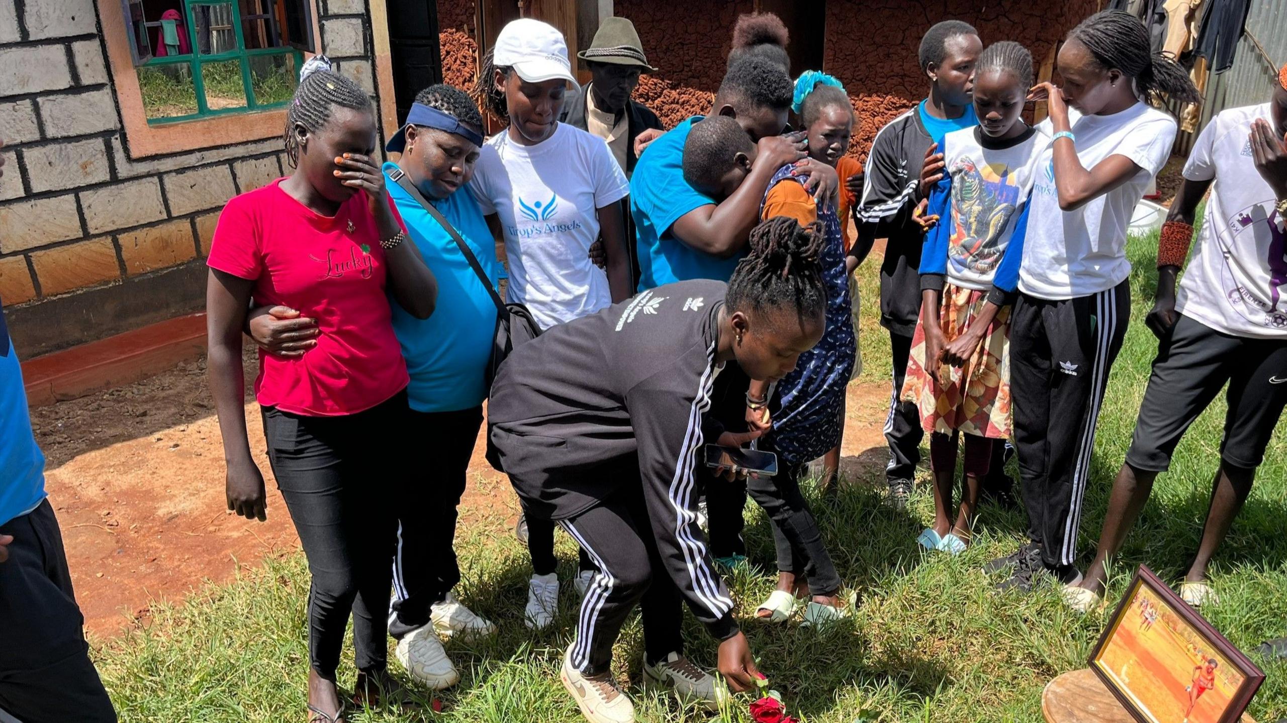 A group of people cry and embrace one another as one lays flowers at the site beside a picture of Rebecca Cheptegei