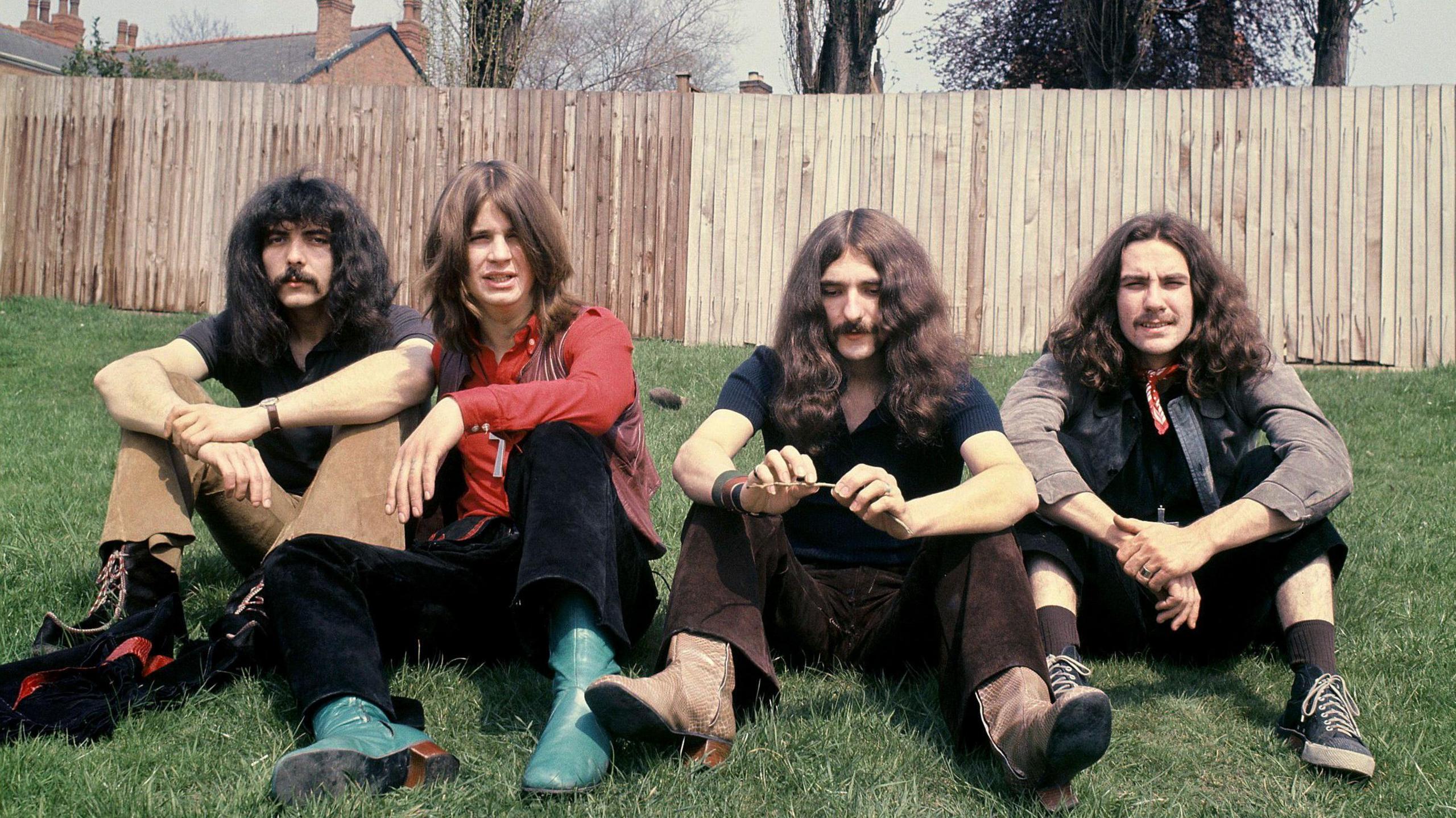 Black Sabbath members ony Iommi, Ozzy Osbourne, Geezer Butler and Bill Ward sat on grass in front of a fence 