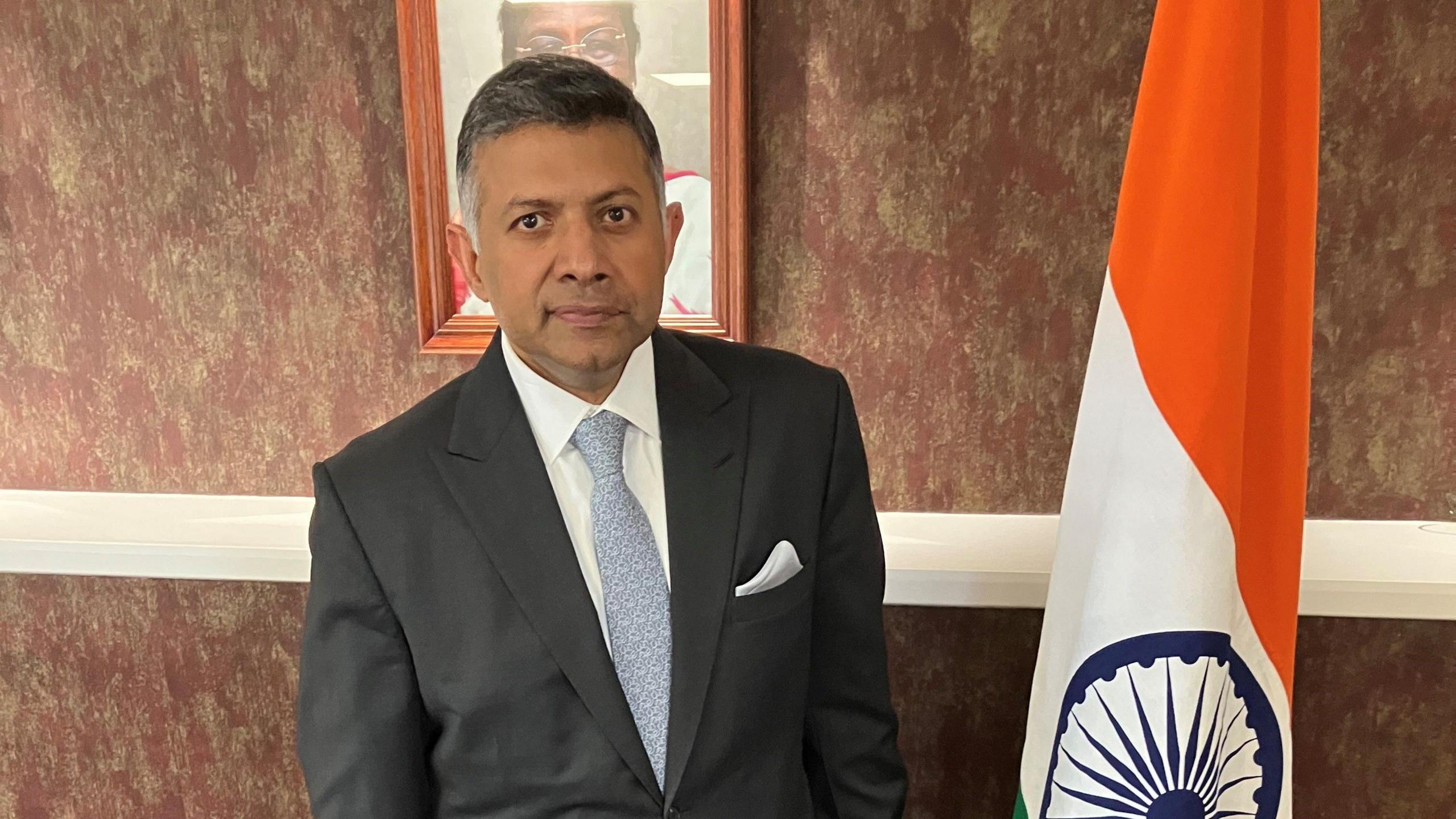 Vikram Doraiswami, High Commissioner of India to the United Kingdom. He is wearing a grey suit and standing next to an Indian flag.