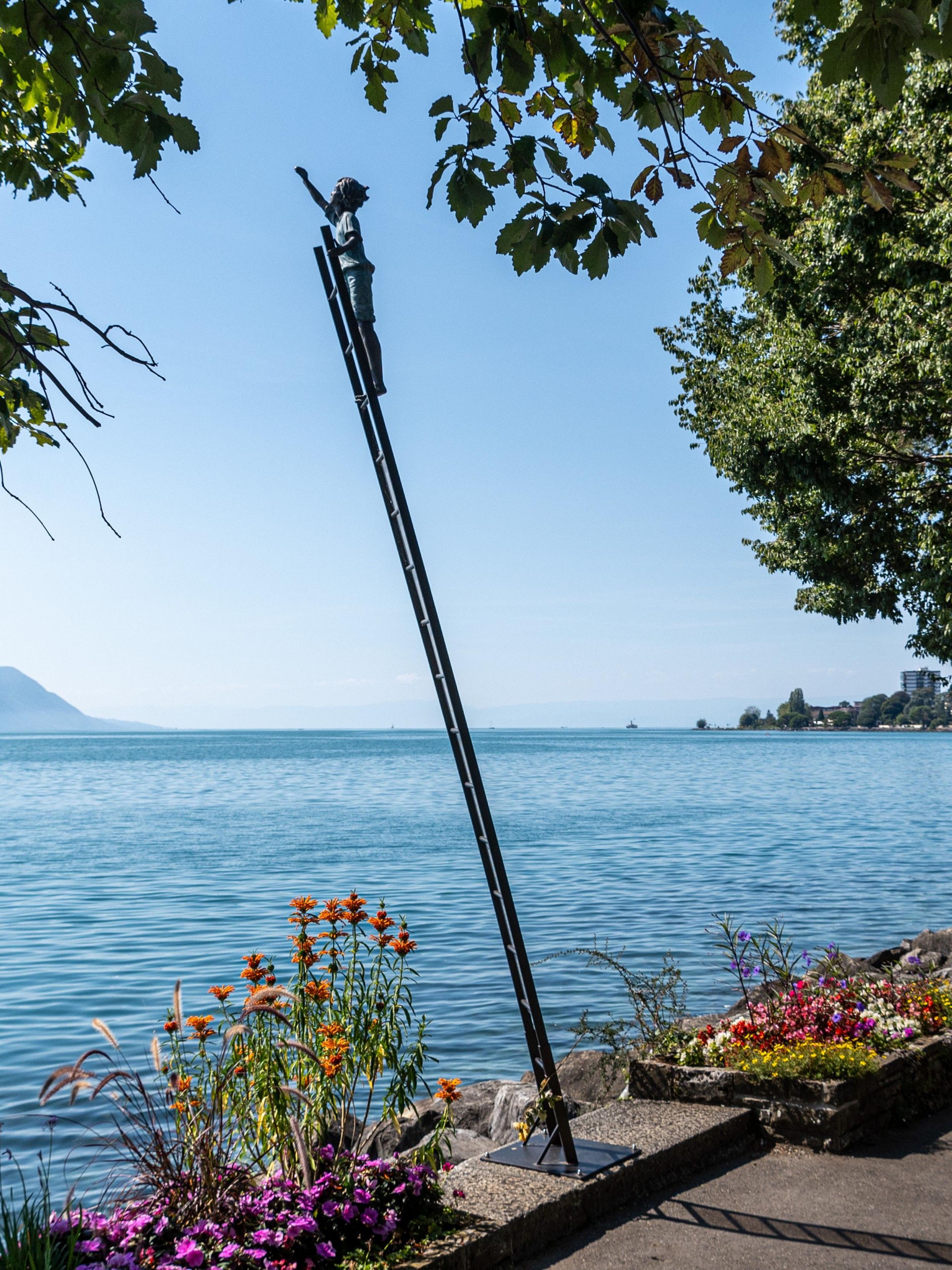 Sculpture of figure reaching out from the top of a ladder next to a lake