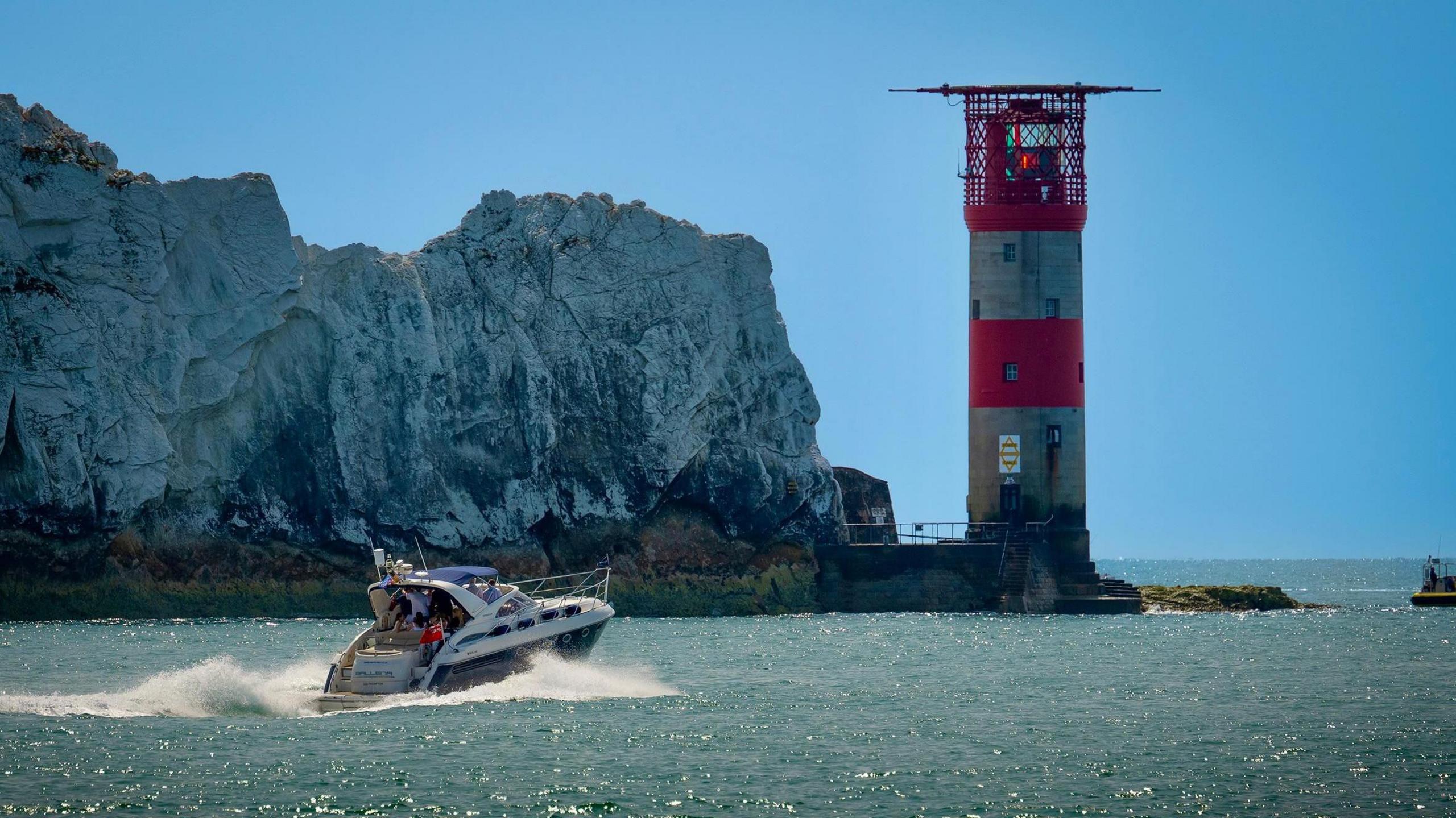 The red and white stripped Needles lighthouse stands at the end of the Needles cliffs off the Isle of Wight Coast. The sea is a bright blue and the blue sky behind is cloudless. The cliffs are a white-grey colour and in the foreground is a white and blue motor cruiser with a white wake sailing towards the lighthouse. 