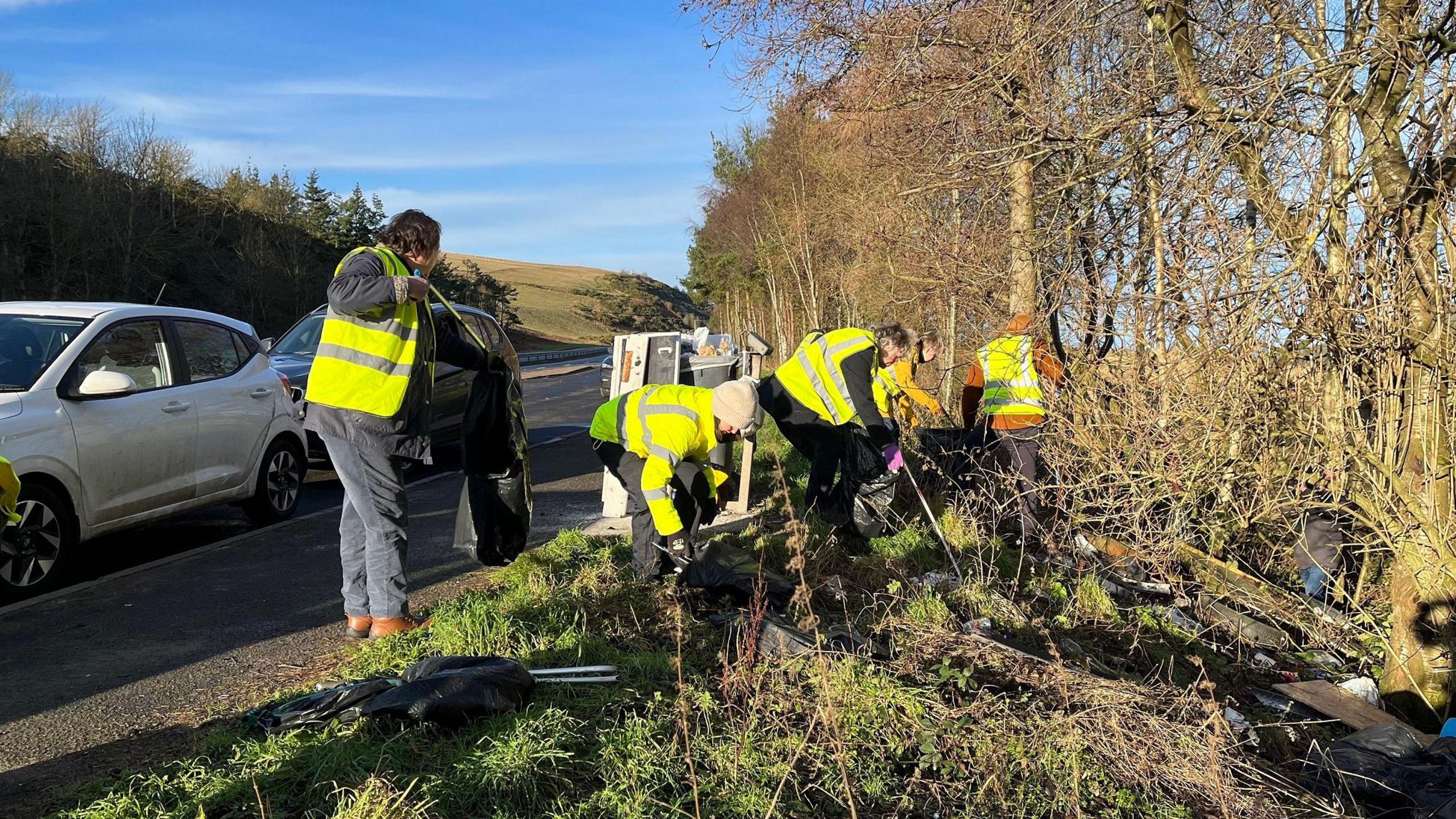 People in high visibility jackets gather litter from a banking with cars and a road in the background