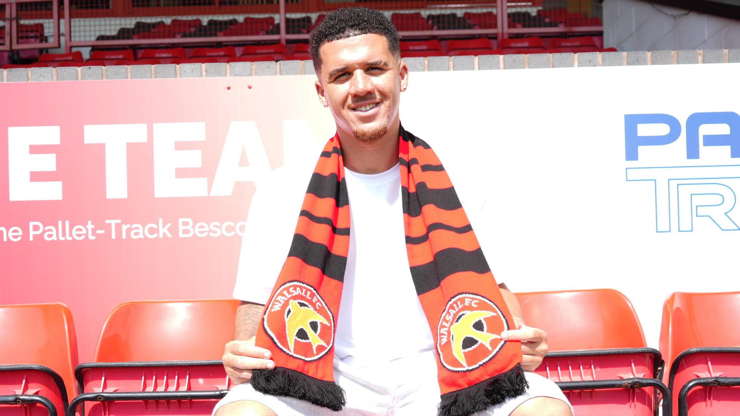 Rico Browne is sitting in the stands at Pallet-Track Stadium with a Walsall scarf around his neck. He is wearing a white tee shirt and shorts. There are red empty seats next to him on either side.