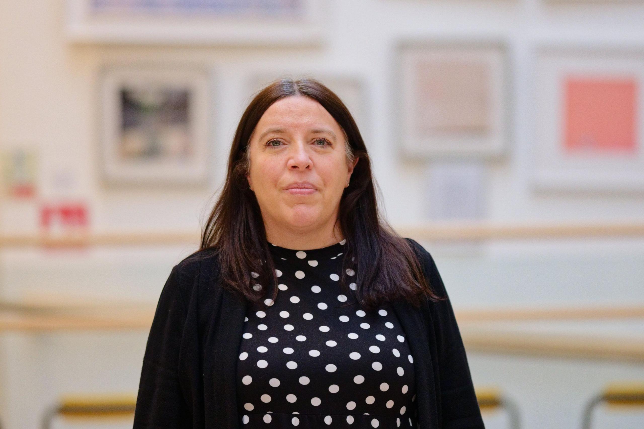 Dawn Bennett is standing in front of the camera wearing a polka dot dress and a navy jumper. 