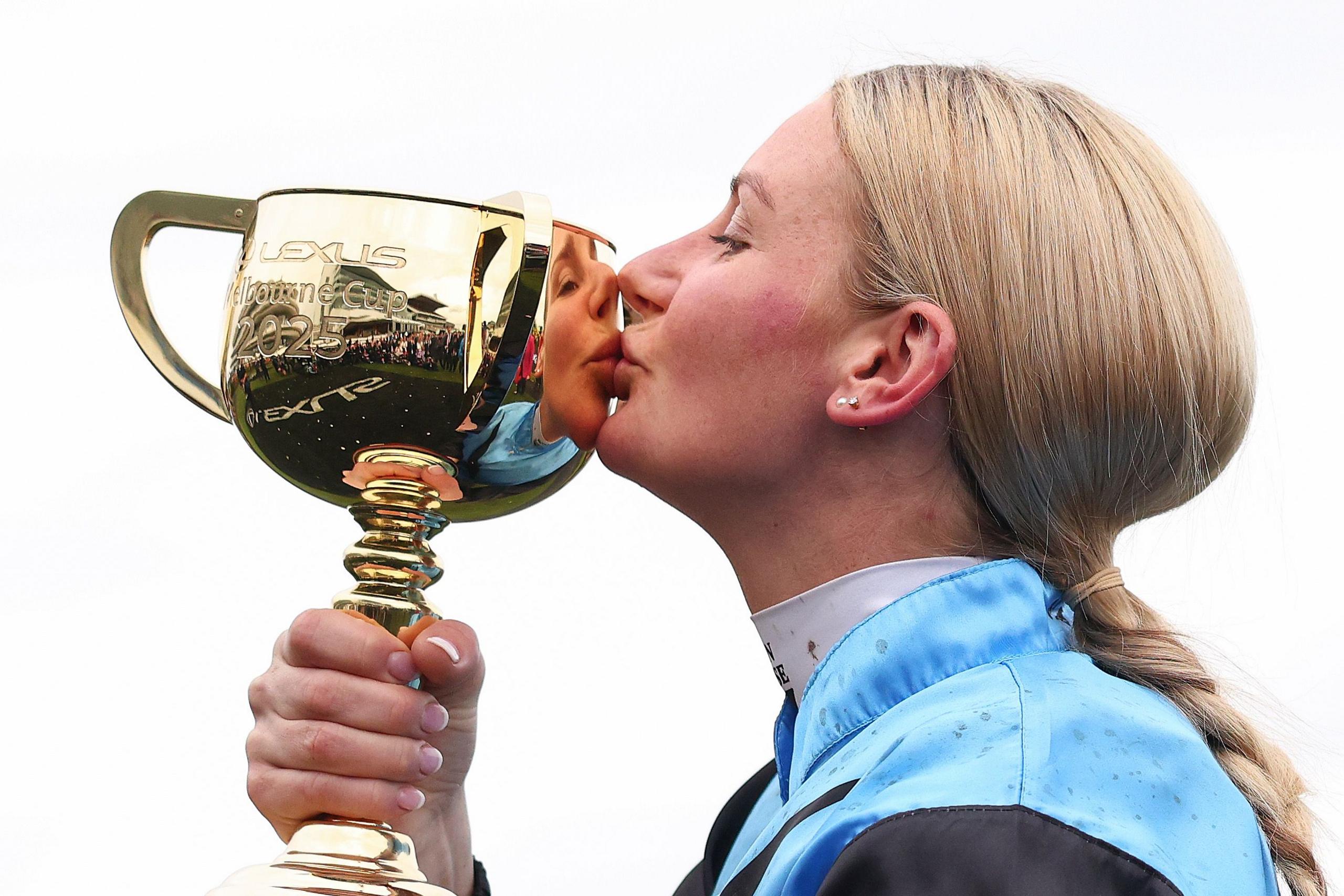 Person wearing a light blue and black racing uniform holding and kissing a large gold trophy engraved with ‘Lexus Melbourne Cup 2025,’ against a plain background, highlighting the shiny metallic surface and detailed handles of the cup.