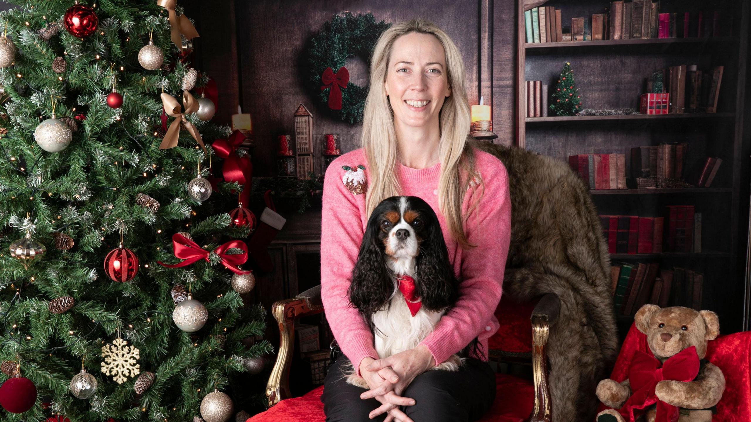 Nicky Crowe, a woman wearing a pink jumper with a Cavalier King Charles Spaniel on her lap. They are sitting next to a Christmas tree.