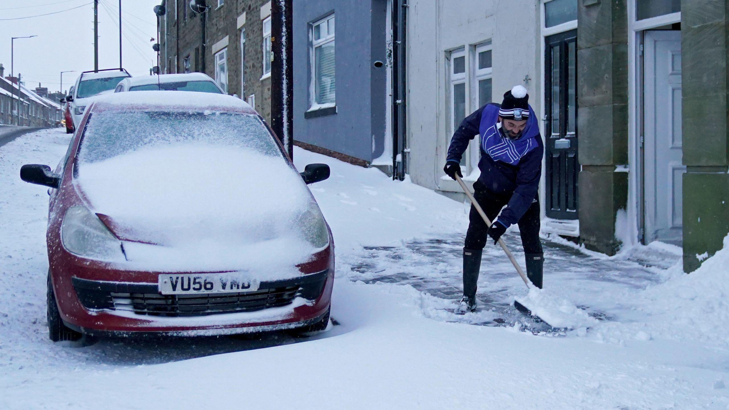 A man shovels snow outside his front door in County Durham. He is dressed in warm clothes and a bobble hat. His home is one of a number of terraced houses on the street. Snow covers the road, pavement and a red car parked outside.